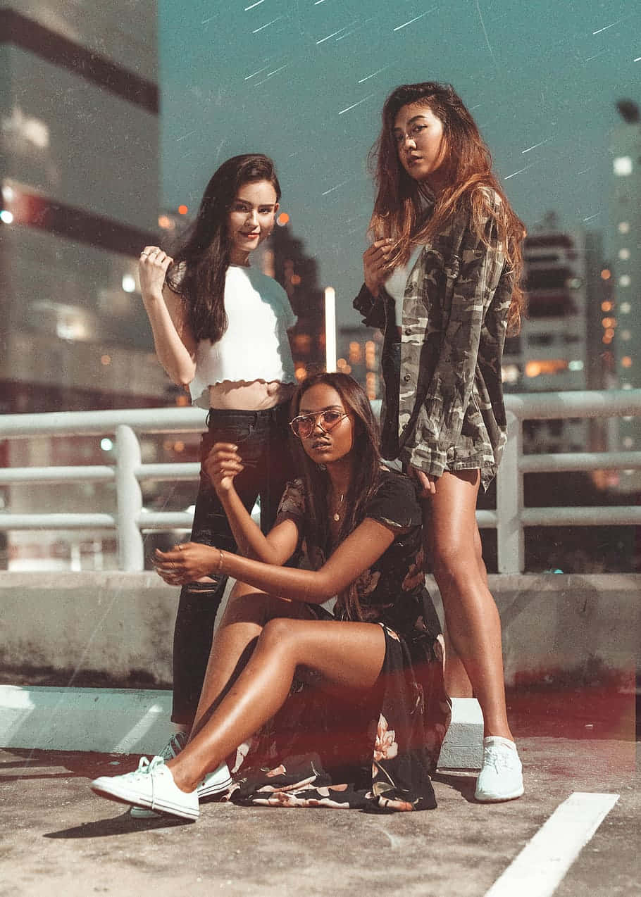 gorgeous 桌布 壁紙 — 三位時尚年輕女性在夜晚的屋頂上自信地擺姿勢，背後是模糊的城市景觀閃爍著光芒。