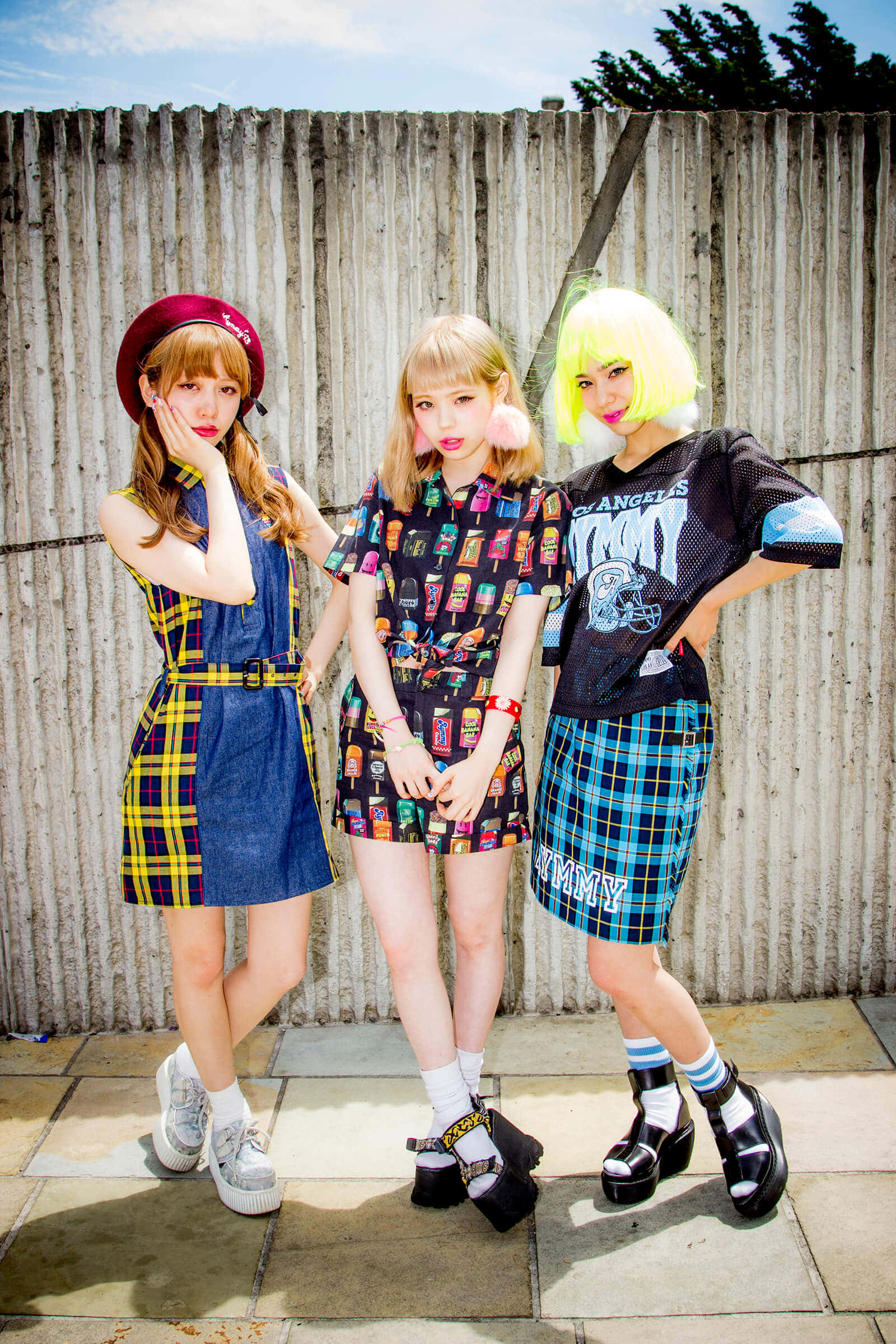 Harajuku Fashion 桌布 壁紙 — 三位時尚年輕女性穿著大膽色彩繽紛的服裝和獨特髮型，在明亮的陽光下自信地靠在風化的木柵欄上擺姿勢。
