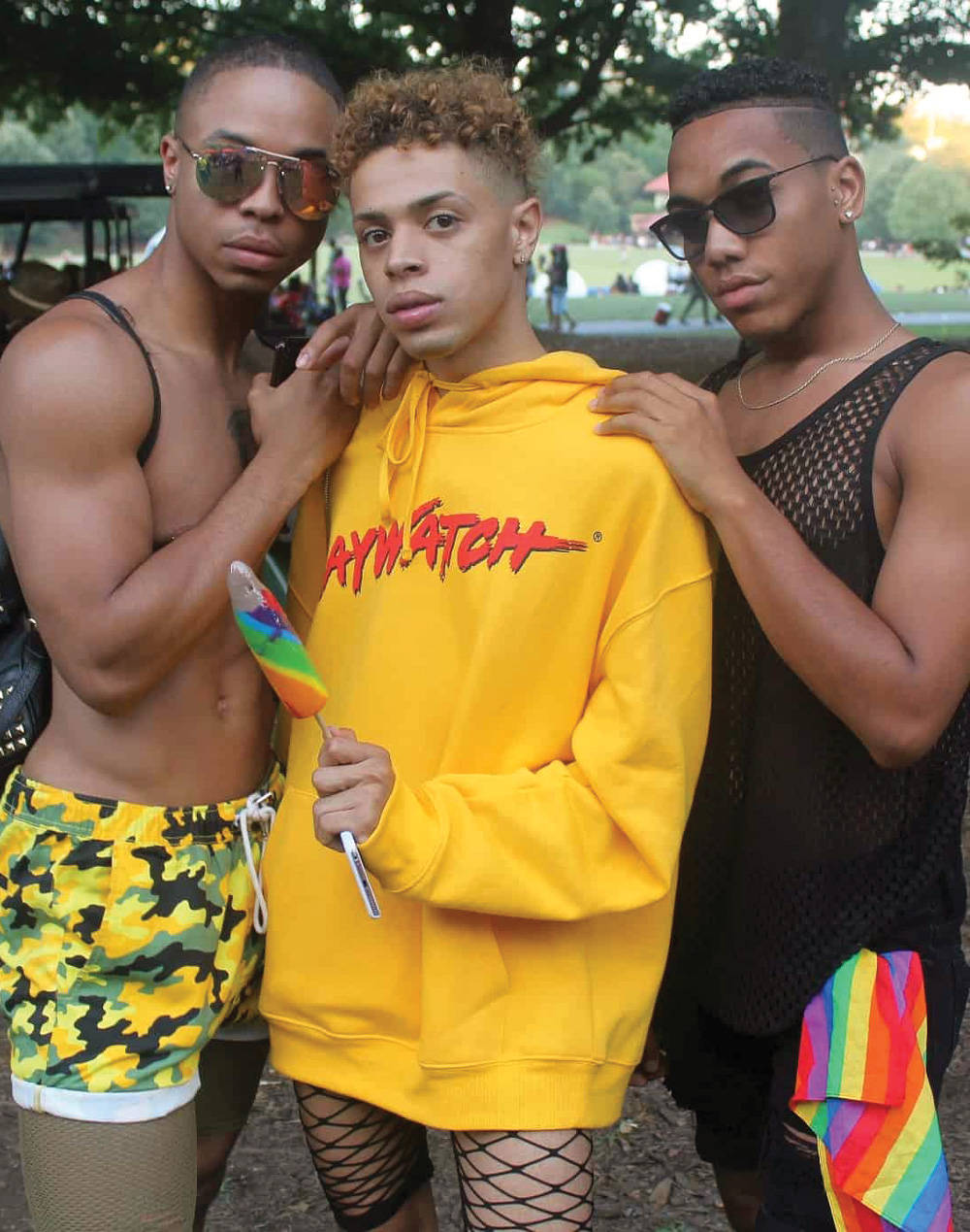 Queer 桌布 壁紙 — 三位時尚年輕男子在公園自信擺姿勢，一人身穿亮黃色連帽衫，手持彩虹棒棒糖，另外兩人以大膽服裝站在他兩側。