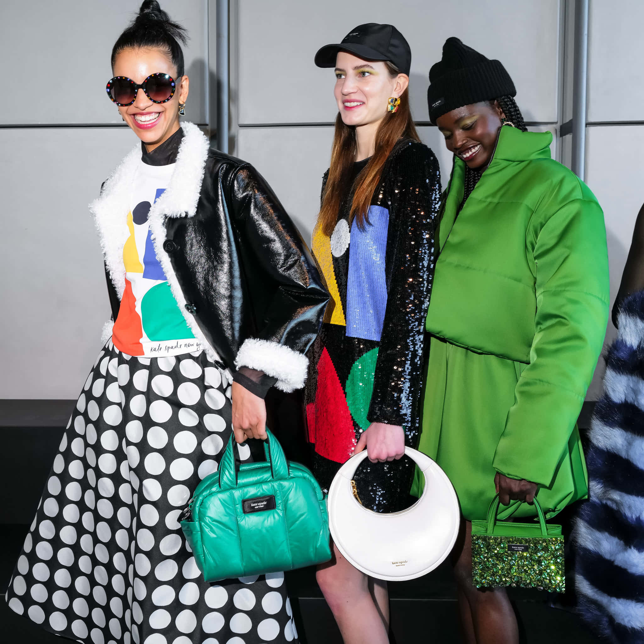 Kate Spade 桌布 壁紙 — 三位時尚模特兒在極簡灰色背景下自信地擺姿勢，穿著大膽時尚服裝，手持色彩繽紛的手袋。