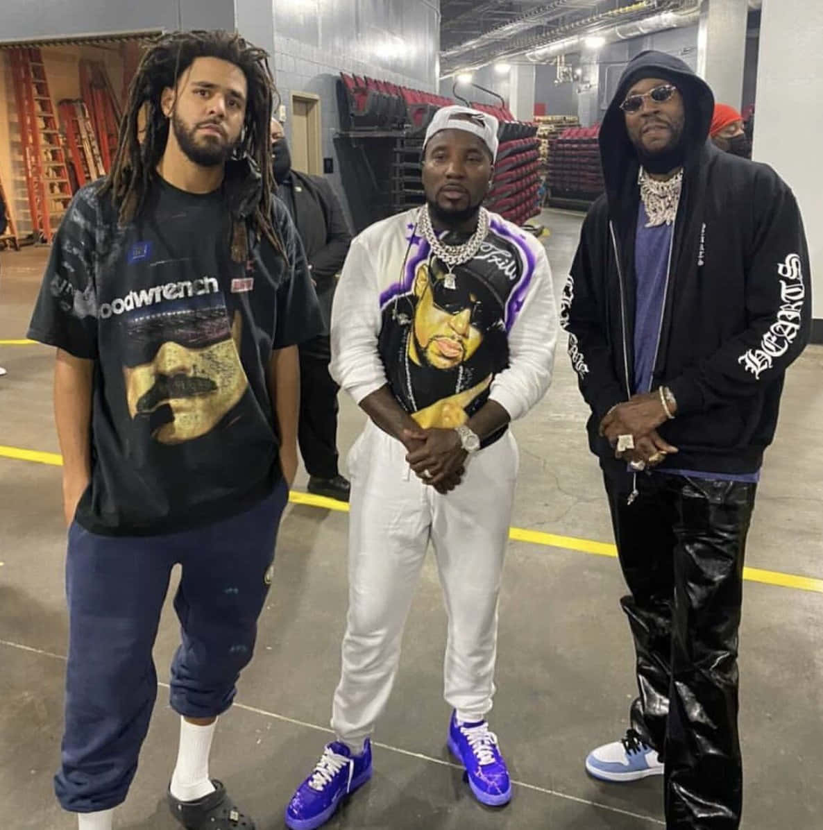2 Chainz 桌布 壁紙 — 三位時尚男士穿著大膽街頭服飾，在後台倉庫中自信擺姿勢，背景有堆疊的紅色椅子和工業照明。