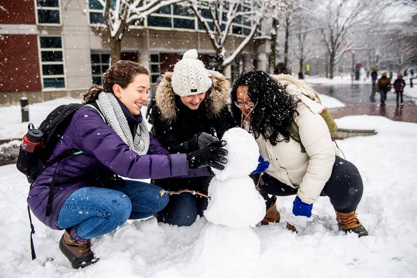 雪 桌布 壁紙 — 三位朋友在白雪覆蓋的校園庭院中，在輕柔的降雪中一起堆雪人，歡聲笑語。