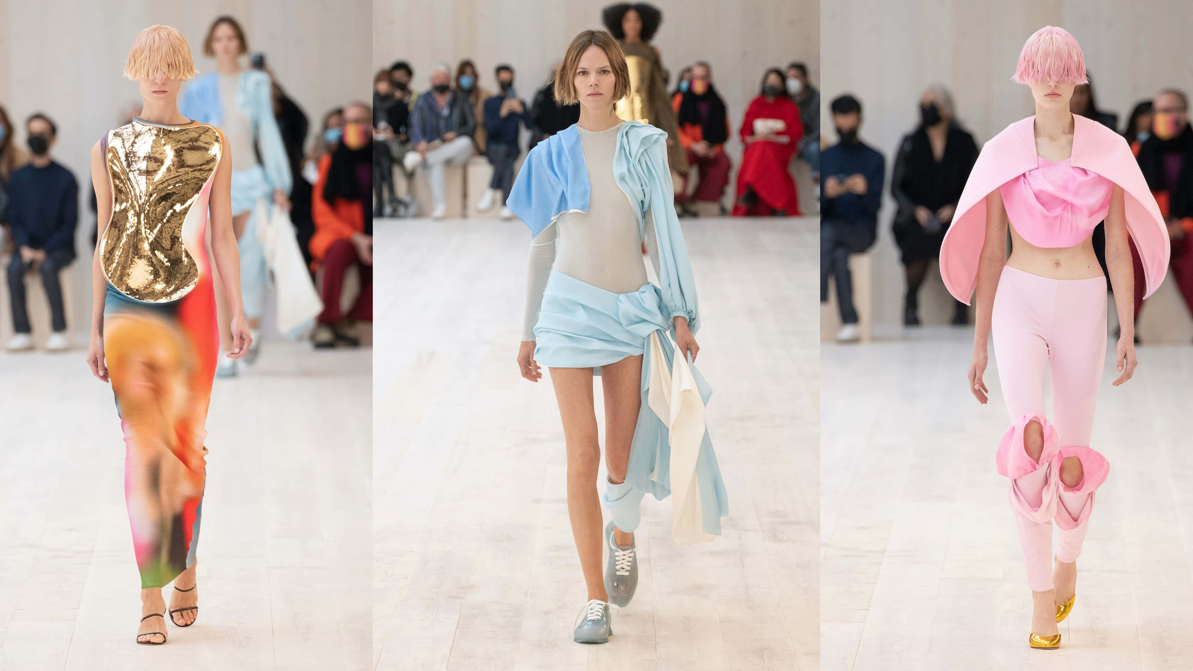 Runway 桌布 壁紙 — 三位模特兒在明亮的時裝伸展台上走秀，展示前衛服裝，包括金屬金色、粉藍色和粉紅色荷葉邊套裝。