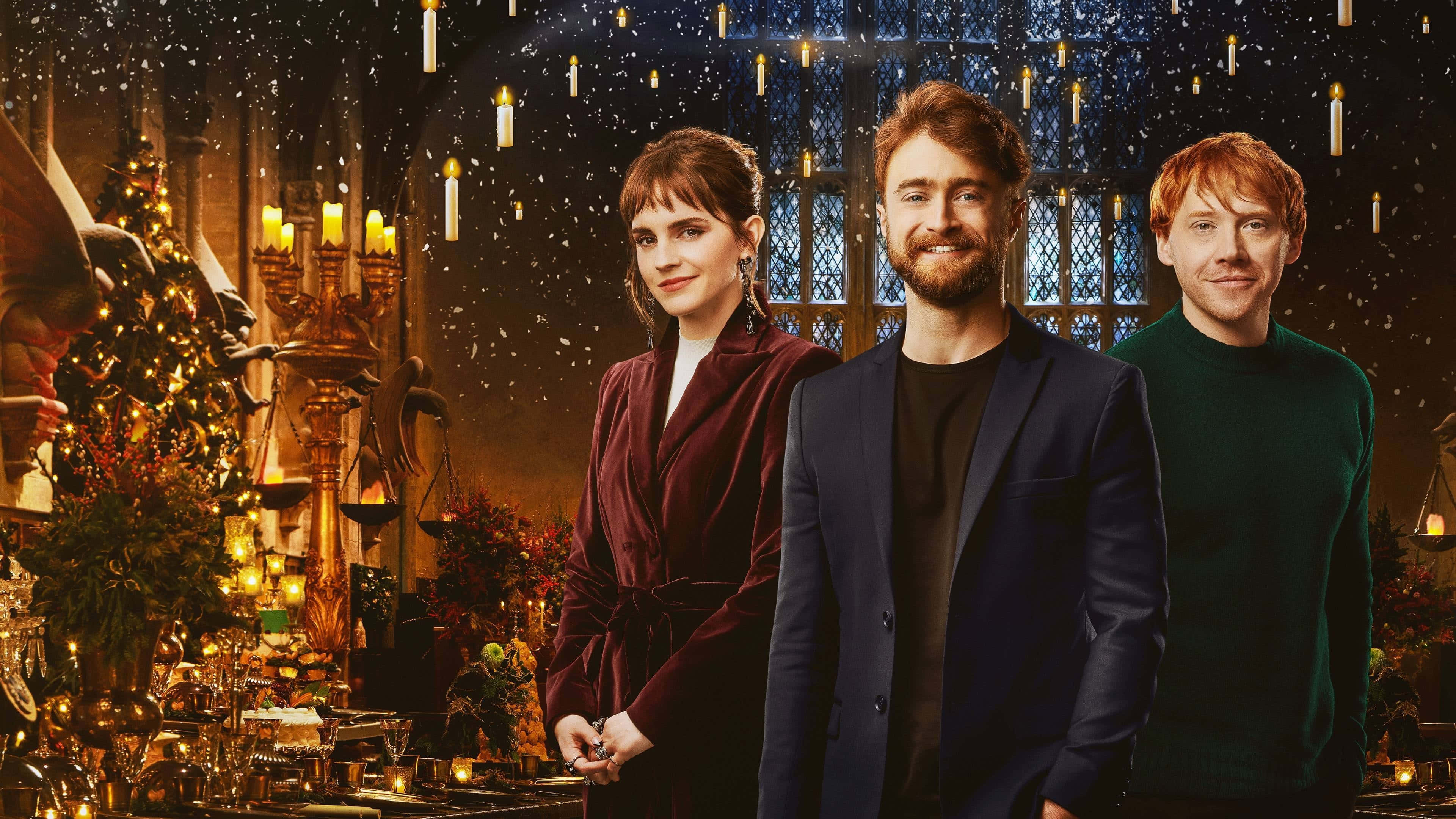 Christmas At Hogwarts 桌布 壁紙 — 三位演員站在一個溫暖燈光、蠟燭裝飾的魔法房間，雪花飄落，節日裝飾點綴其間。