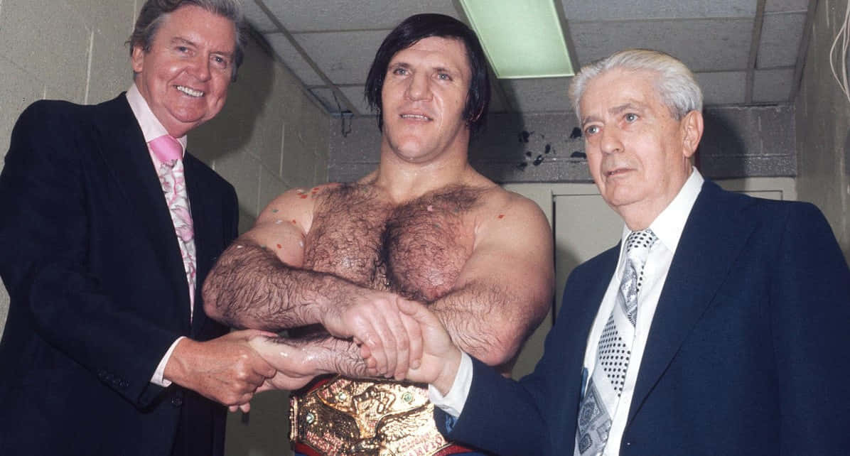WWE 桌布 壁紙 — 三位男士在後台走廊，一位肌肉發達的摔角手在兩位西裝男士之間手持冠軍腰帶。