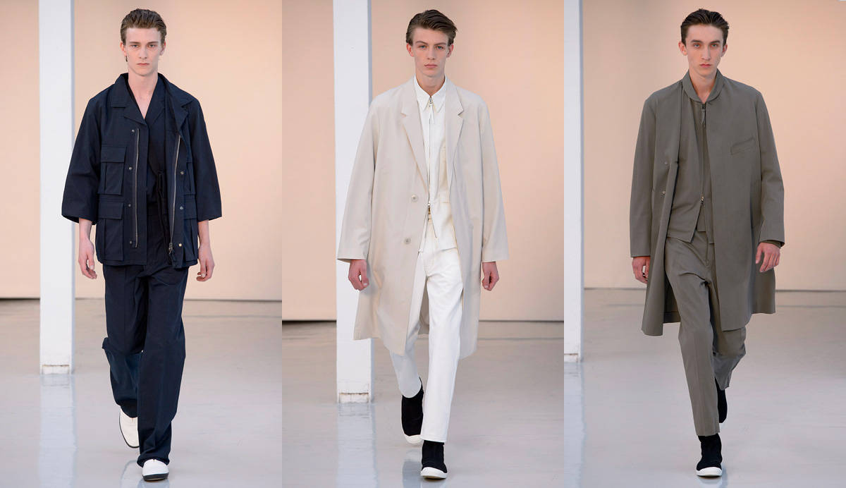 Mens Fashion 桌布 壁紙 — 三位男模特兒自信地走在簡約的伸展台上，穿著海軍藍、米色和灰色調的前衛訂製服裝。