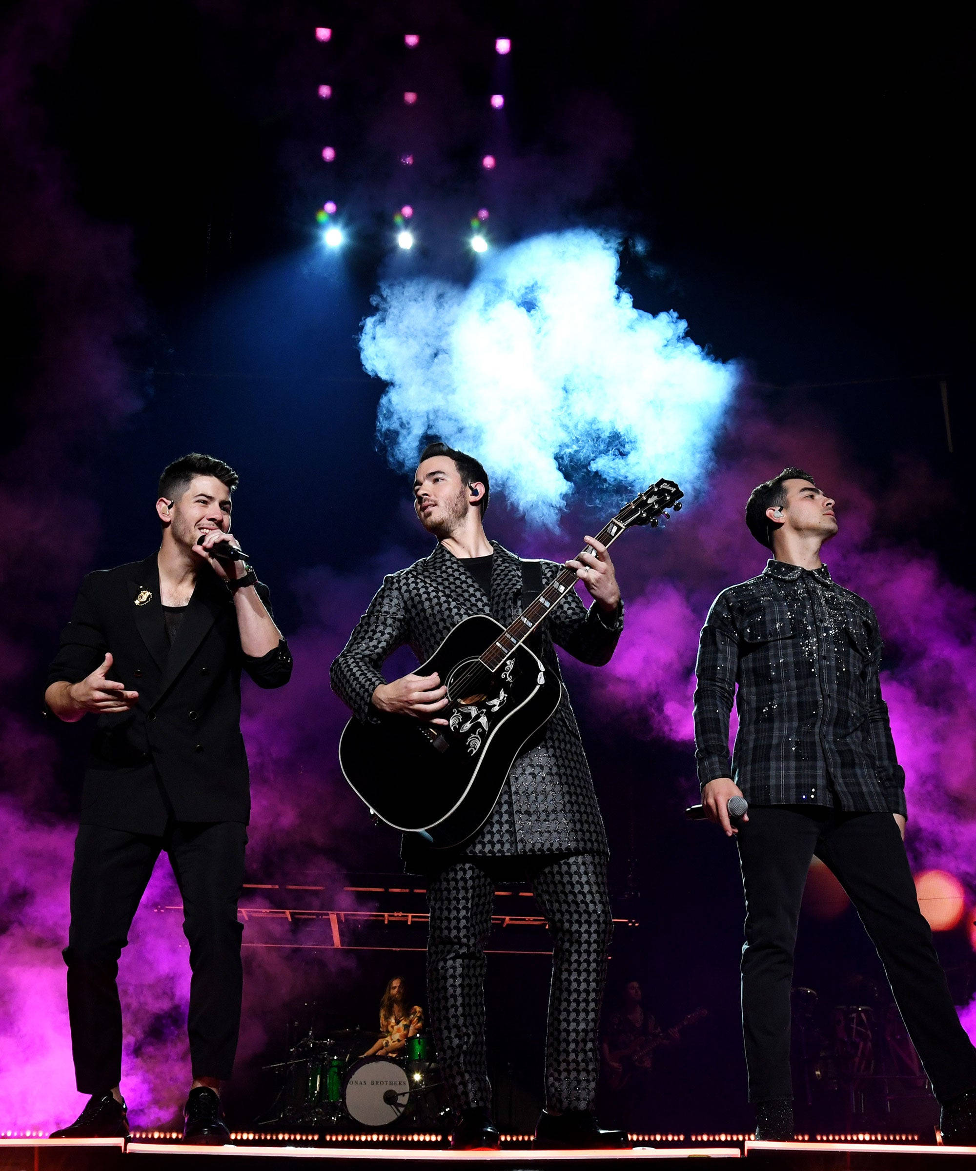 Jonas Brothers 桌布 壁紙 — 三位男音樂家在充滿紫色煙霧和藍色聚光燈的活力舞台上表演，其中一位正在彈吉他。