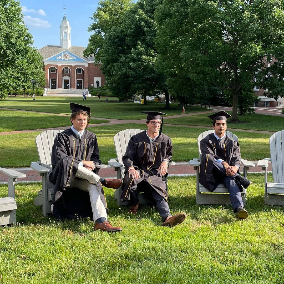 college 桌布 壁紙 — 三位畢業生身穿學士袍和四方帽，輕鬆地坐在陽光普照的校園草坪上的木椅上，背景是歷史建築和茂密的樹木。