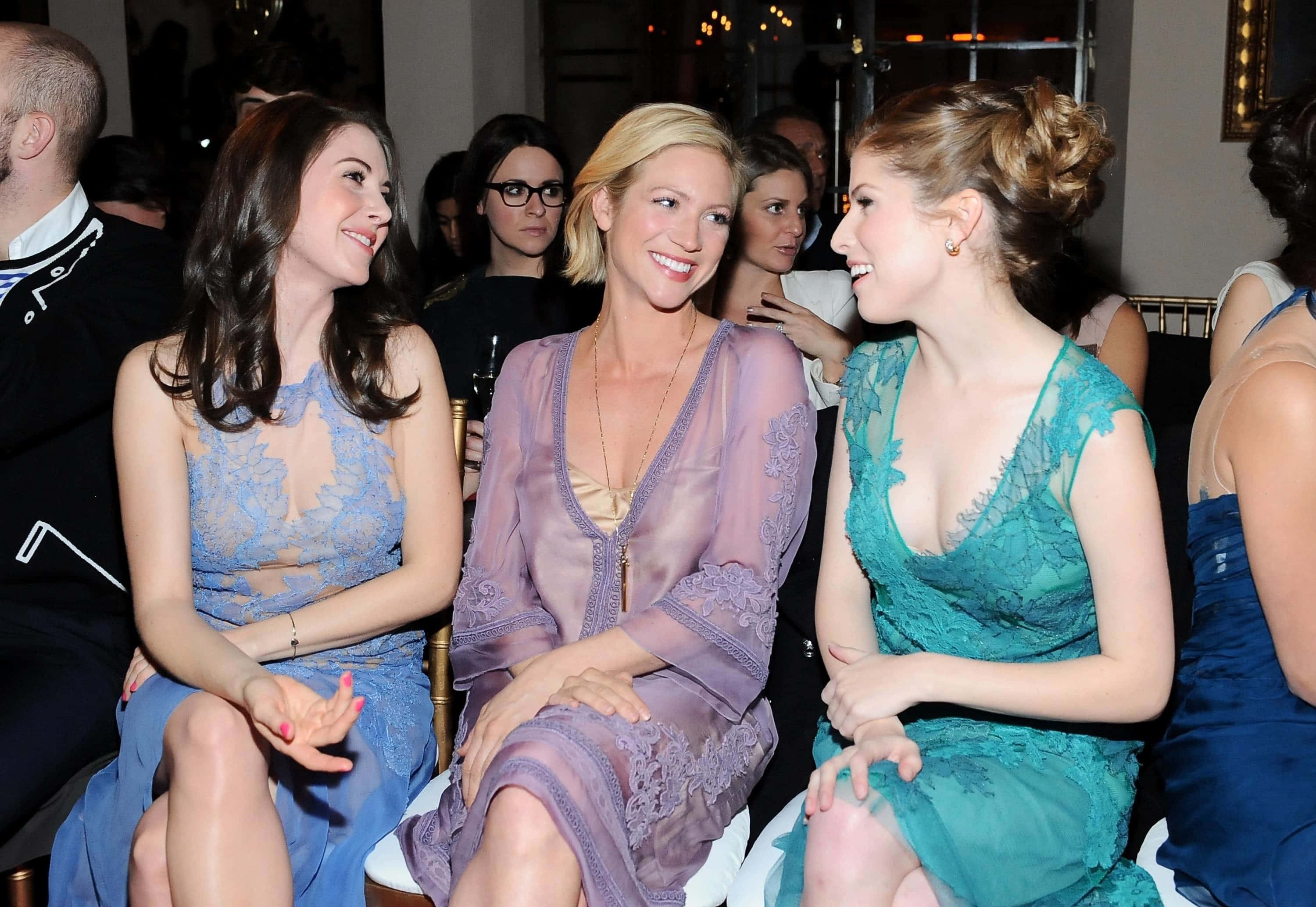 brittany snow 桌布 壁紙 — 三位穿著優雅蕾絲裙的女性在溫暖的環境燈光下，在一個迷人的室內活動中一起大笑。