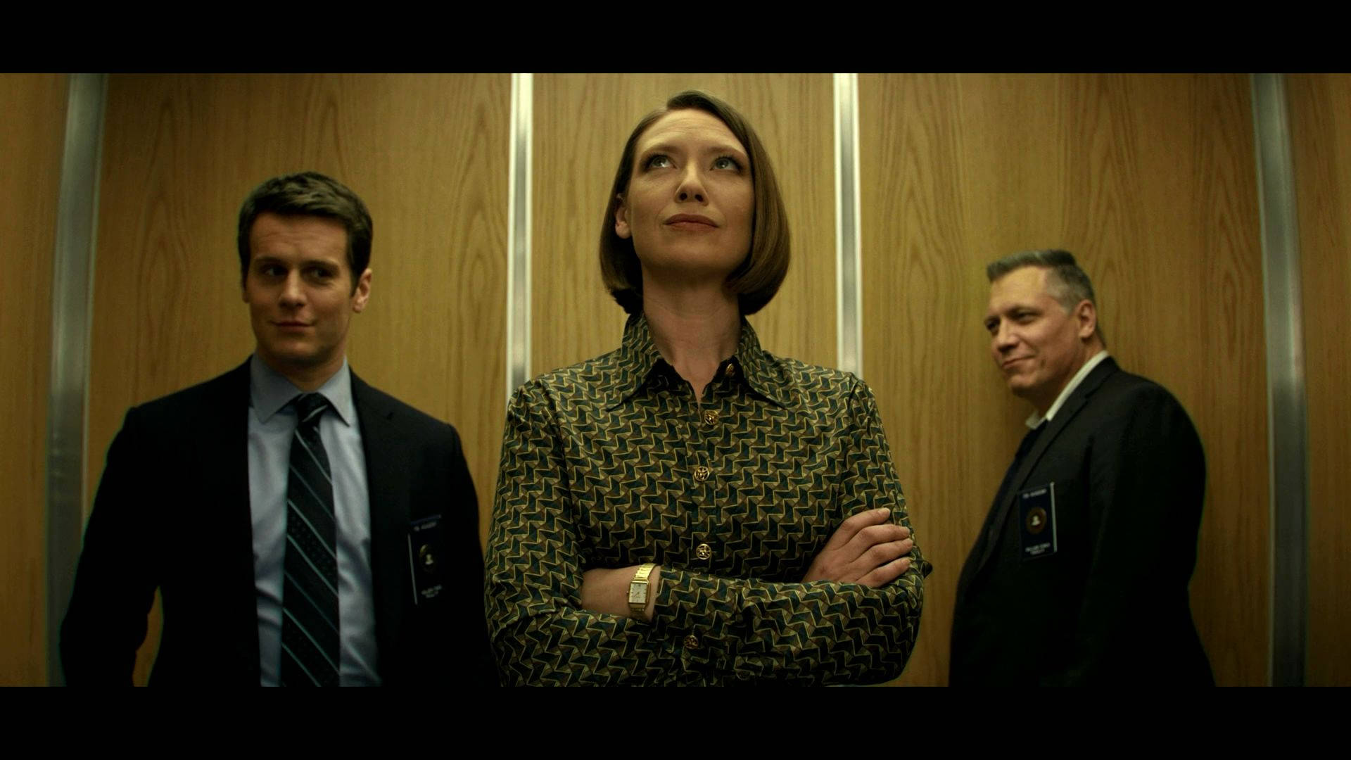 anna torv 桌布 壁紙 — 三位穿著正式服裝的人士站在木質面板的電梯內，表情緊張，雙臂交叉。