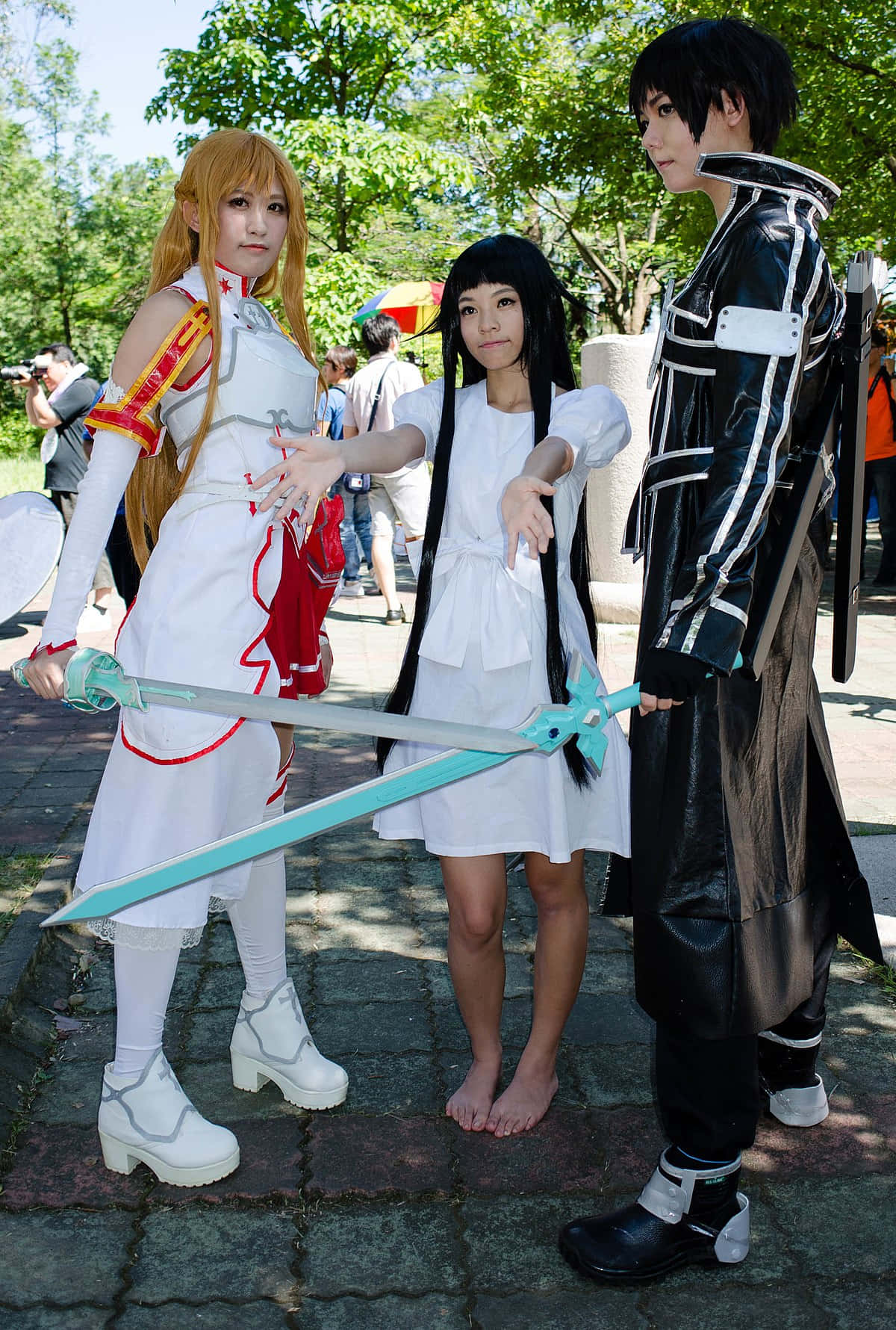 Sword Art Online Cosplay 桌布 壁紙 — 三位穿著精細動漫服裝的角色扮演者在戶外陽光下，共同手持一把發光的劍擺姿勢。