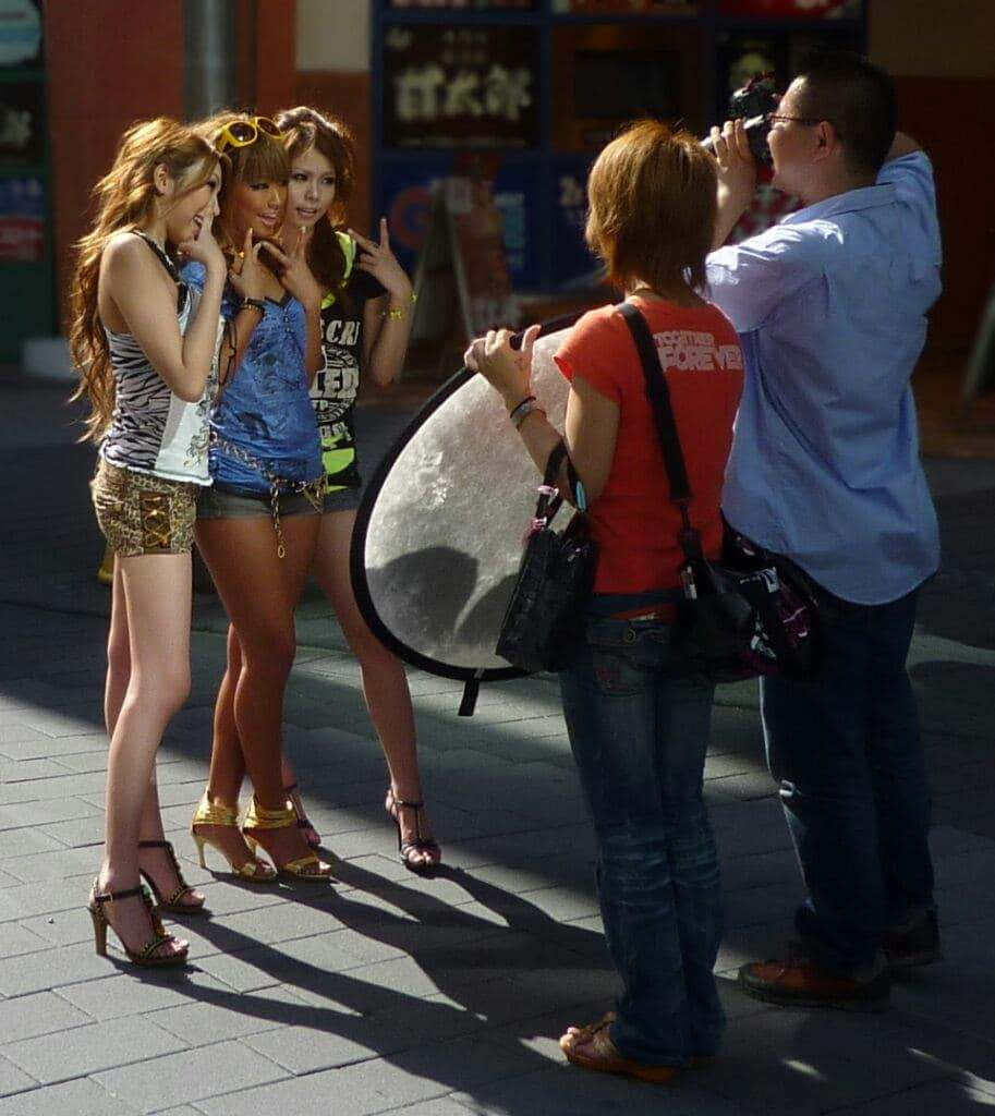 Gyaru Fashion 桌布 壁紙 — 三位穿著高跟鞋的年輕女性在明亮的陽光下為一位手持反光板的街頭攝影師擺姿勢。