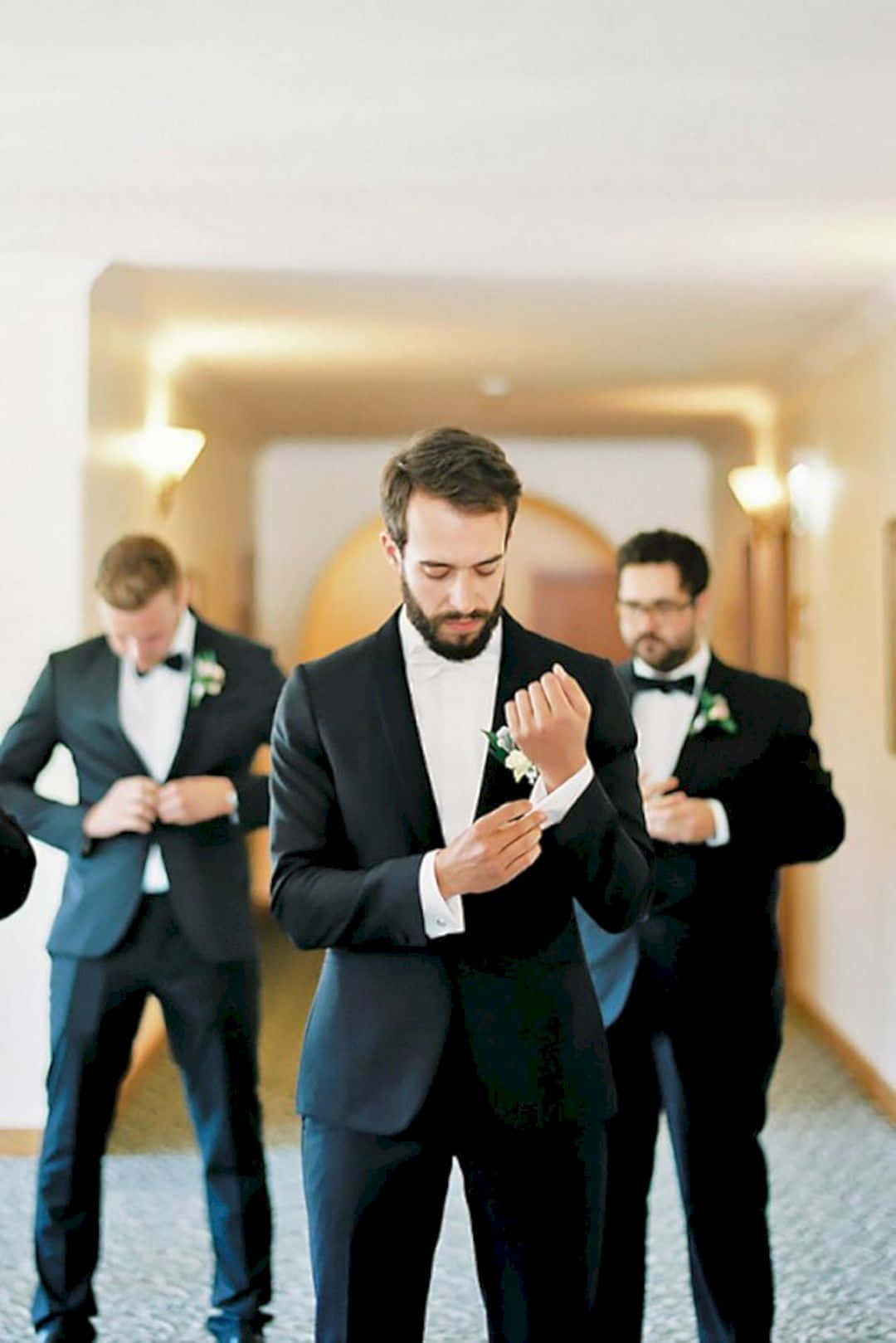 Groomsman 桌布 壁紙 — 三位穿著黑色禮服的伴郎在婚禮前於溫暖燈光的走廊調整他們的蝴蝶結和襟花。