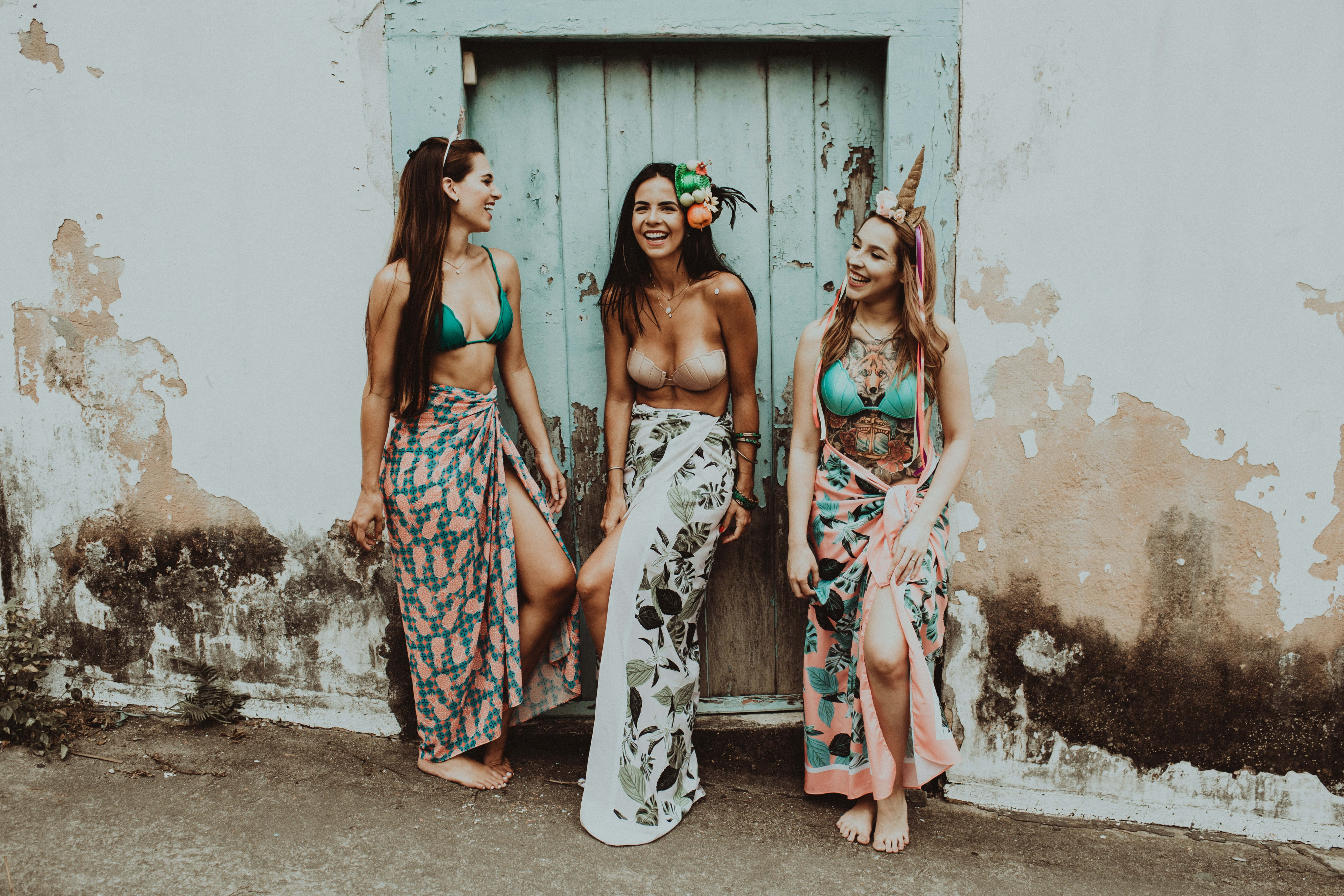 Summer Fashion 桌布 壁紙 — 三位笑容滿面的女士身穿色彩繽紛圖案沙龍和比基尼，在鄉村風格的牆上，靠著一扇褪色的藍色門，俏皮地擺姿勢。