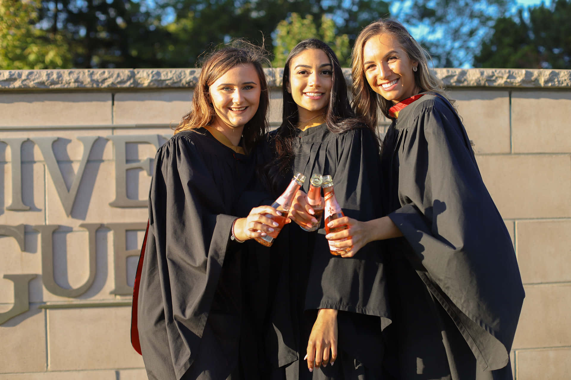 Nurses Graduation 桌布 壁紙 — 三位笑容滿面的畢業生身穿黑色畢業袍，在刻有字母的石牆前用粉紅色飲料乾杯。