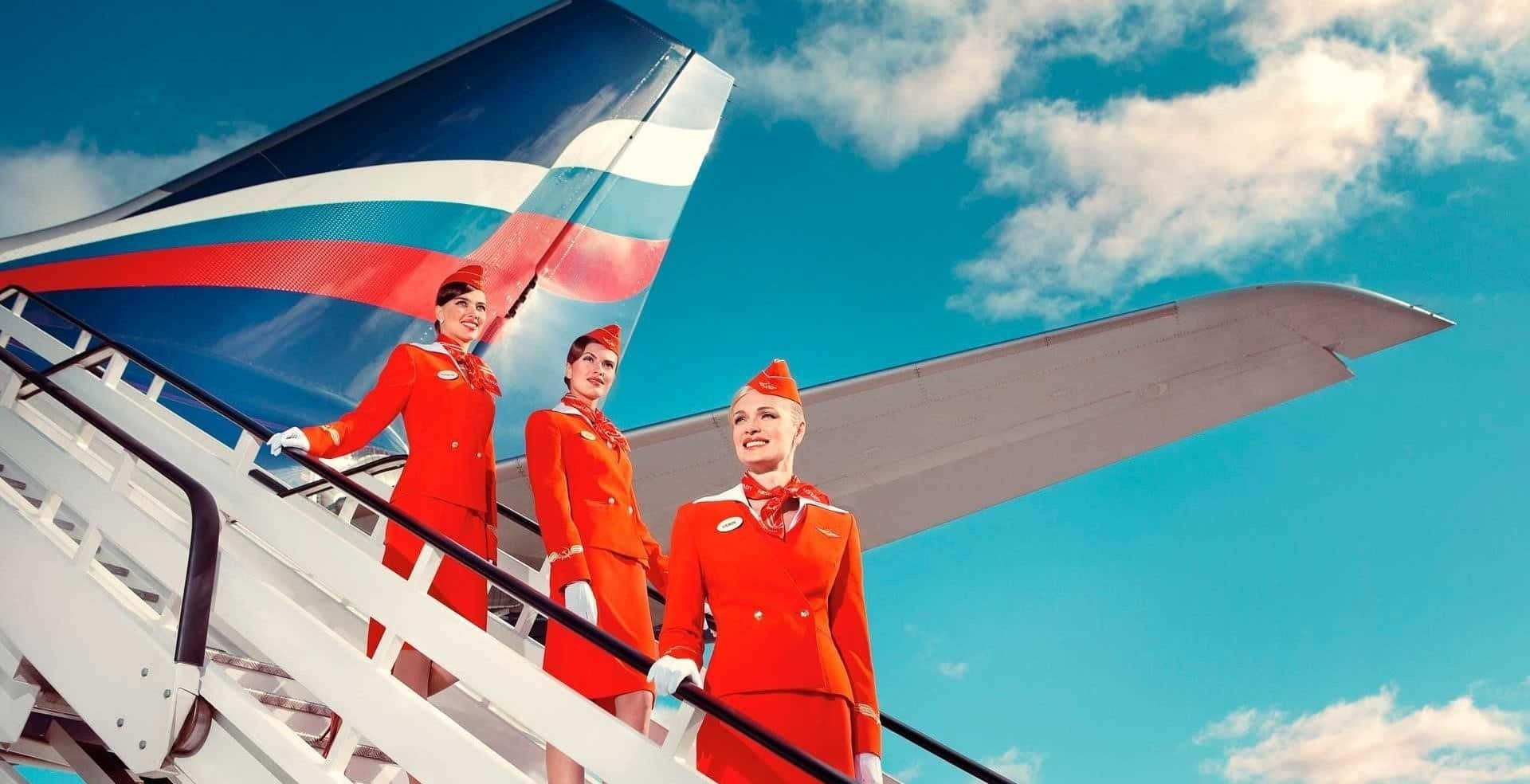 Flight Attendant 桌布 壁紙 — 三位笑容滿面的空服員身穿鮮紅色制服，站在飛機樓梯上，背景是晴朗的藍天和蓬鬆的白雲。