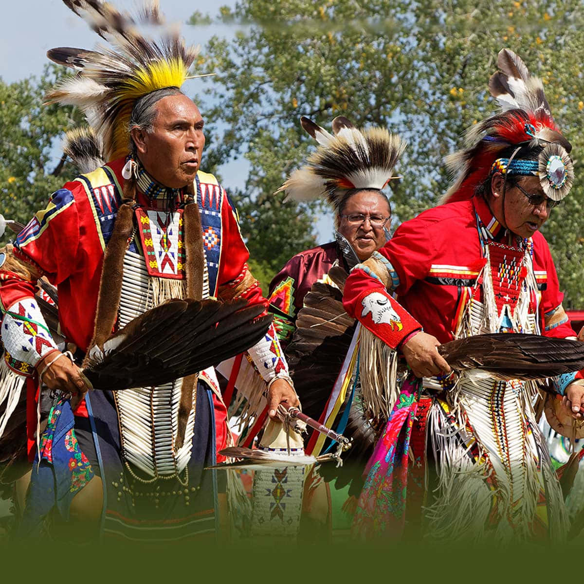 Native American 桌布 壁紙 — 三位美洲原住民男子身穿鮮豔的傳統服飾，頭戴羽毛頭飾，在明亮的陽光下戶外擊鼓跳舞。