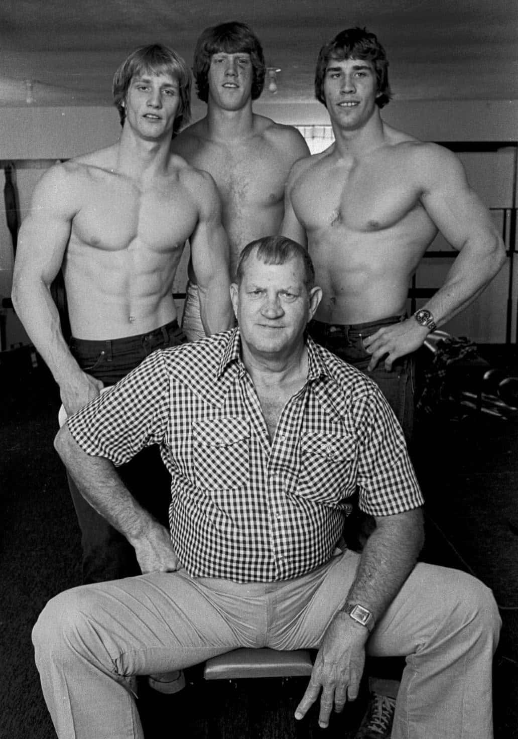 摔角 桌布 壁紙 — 三位肌肉發達、赤裸上身的男士站在一位身穿格紋襯衫的坐著長者後面，背景是健身房。