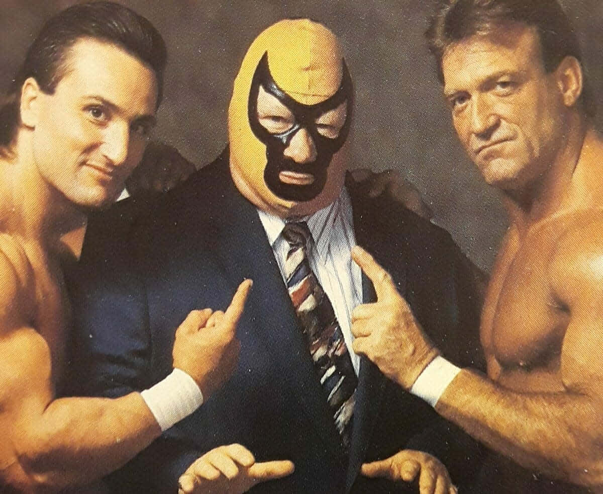 WWE 桌布 壁紙 — 三位肌肉發達的摔角手一起擺姿勢，其中一位戴著黃色面具穿著西裝，而其他人赤裸上身指著他。