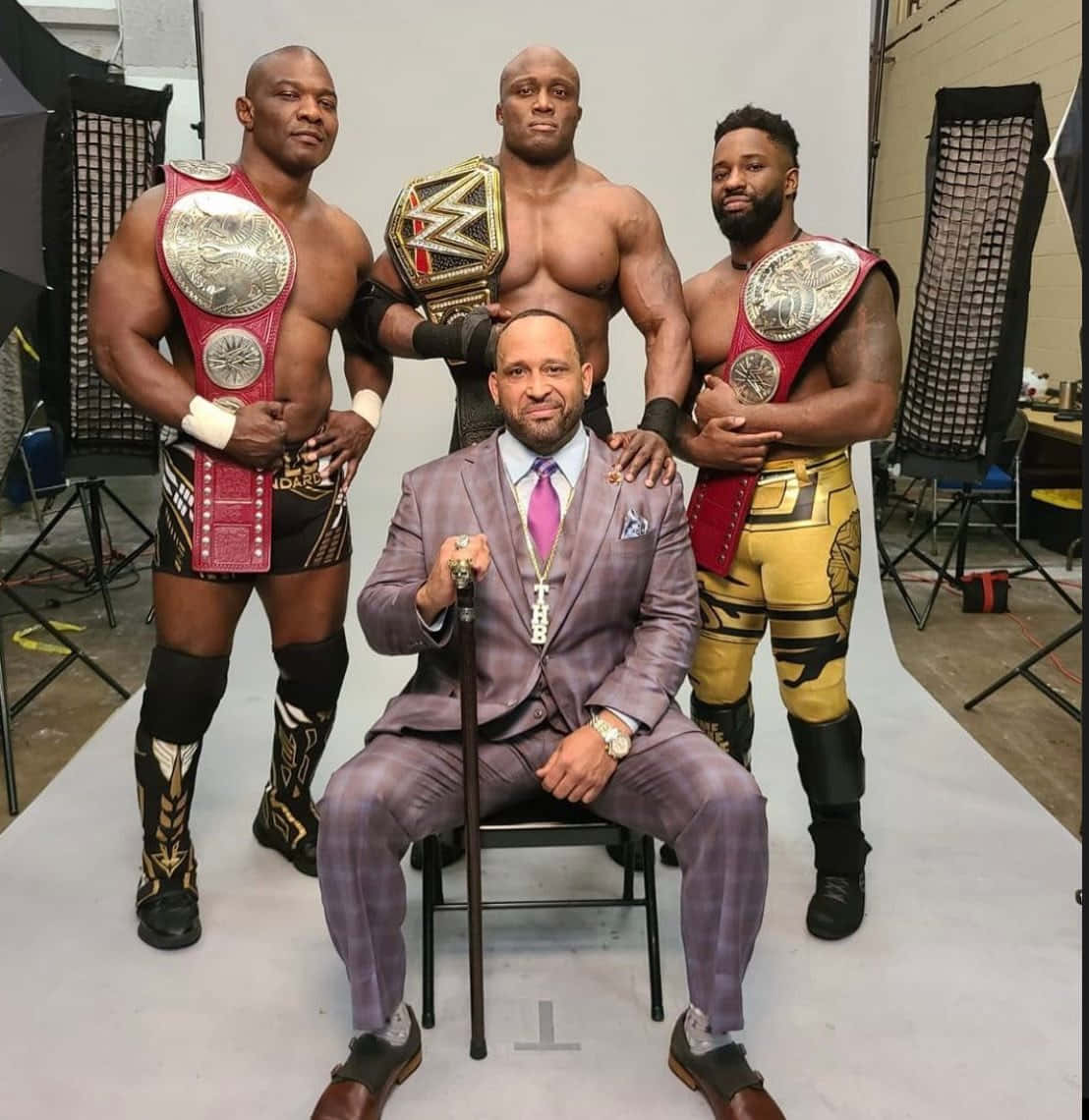 WWE 桌布 壁紙 — 三位肌肉發達的摔角手穿著色彩繽紛的裝備，站在一位身穿紫色西裝、手持枴杖的坐著男子後面，在攝影棚內。