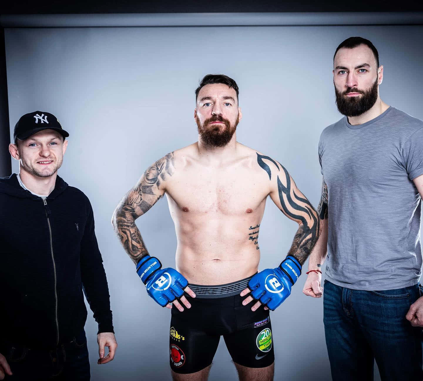 MMA 桌布 壁紙 — 三位肌肉發達的男士在攝影棚自信擺姿勢，其中一位沒穿上衣，戴著藍色手套，刺青在純灰色背景下清晰可見。