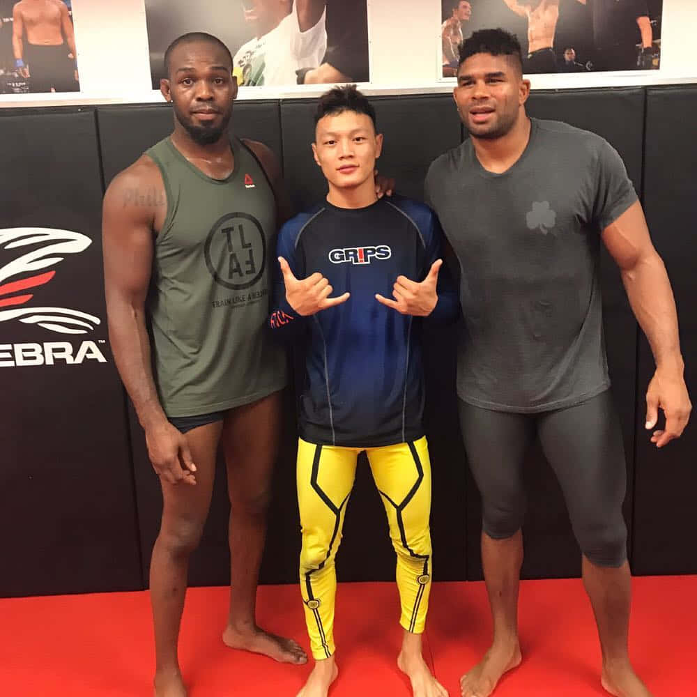 MMA 桌布 壁紙 — 三位肌肉發達的男士在鋪有紅色軟墊和貼有格鬥海報的健身房一起擺姿勢。