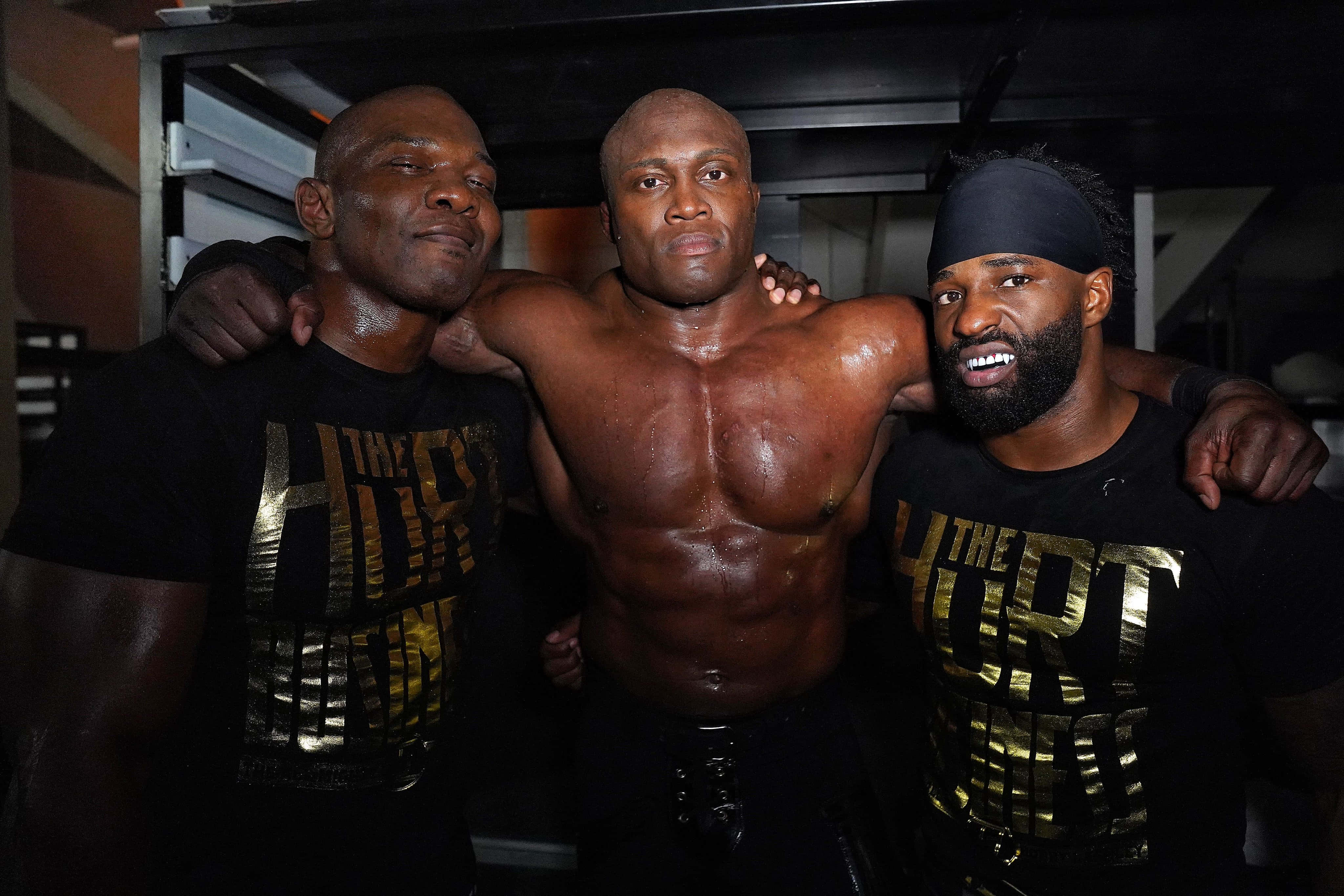 WWE 桌布 壁紙 — 三位肌肉發達的男士身穿黑色「The Hurt Business」T恤，在燈光昏暗的後台區域自信地互相搭肩擺姿勢。