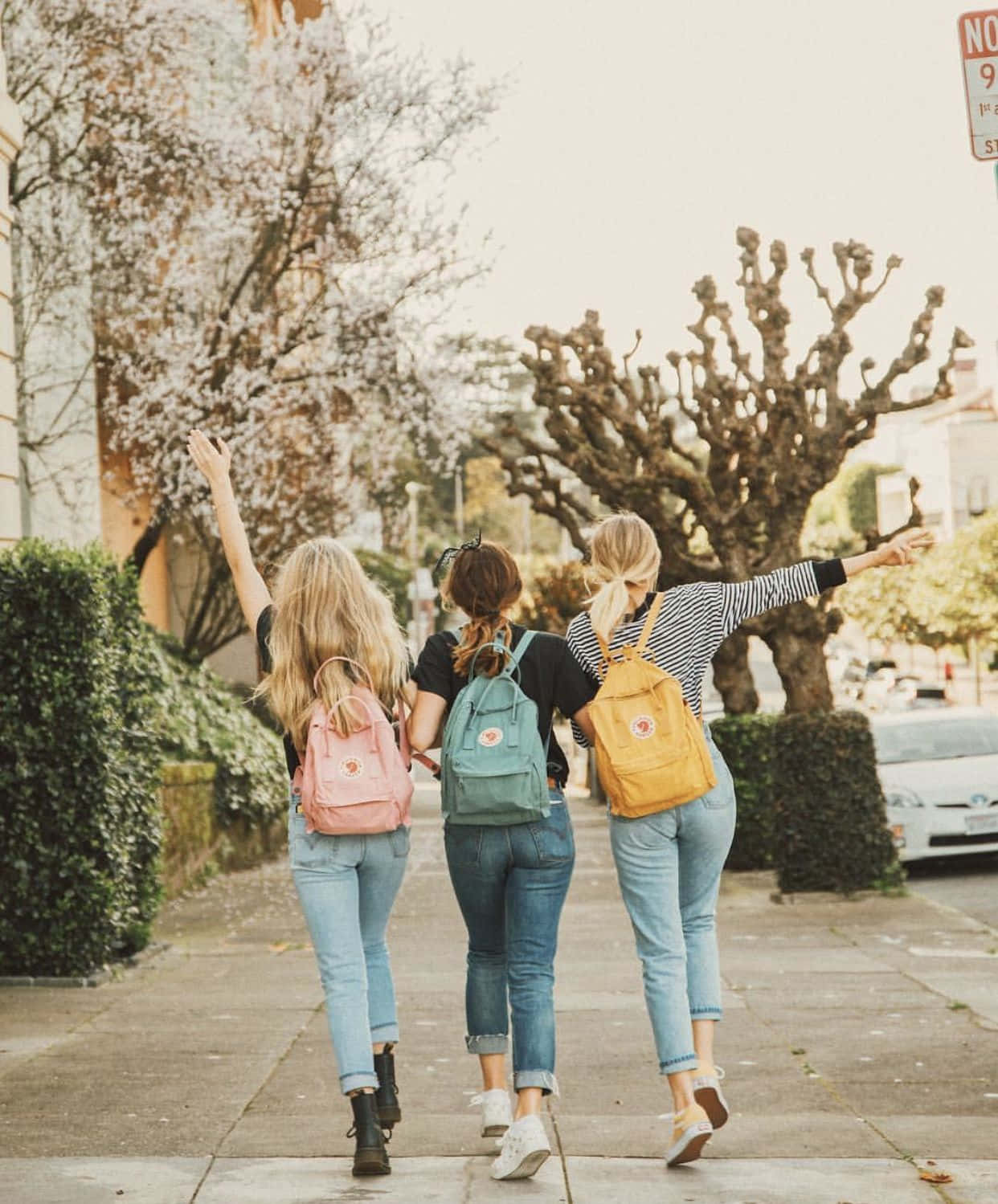 Friends 桌布 壁紙 — 三位背著彩色背包的朋友在綠樹成蔭的人行道上漫步，高舉雙臂，感受快樂的自由。