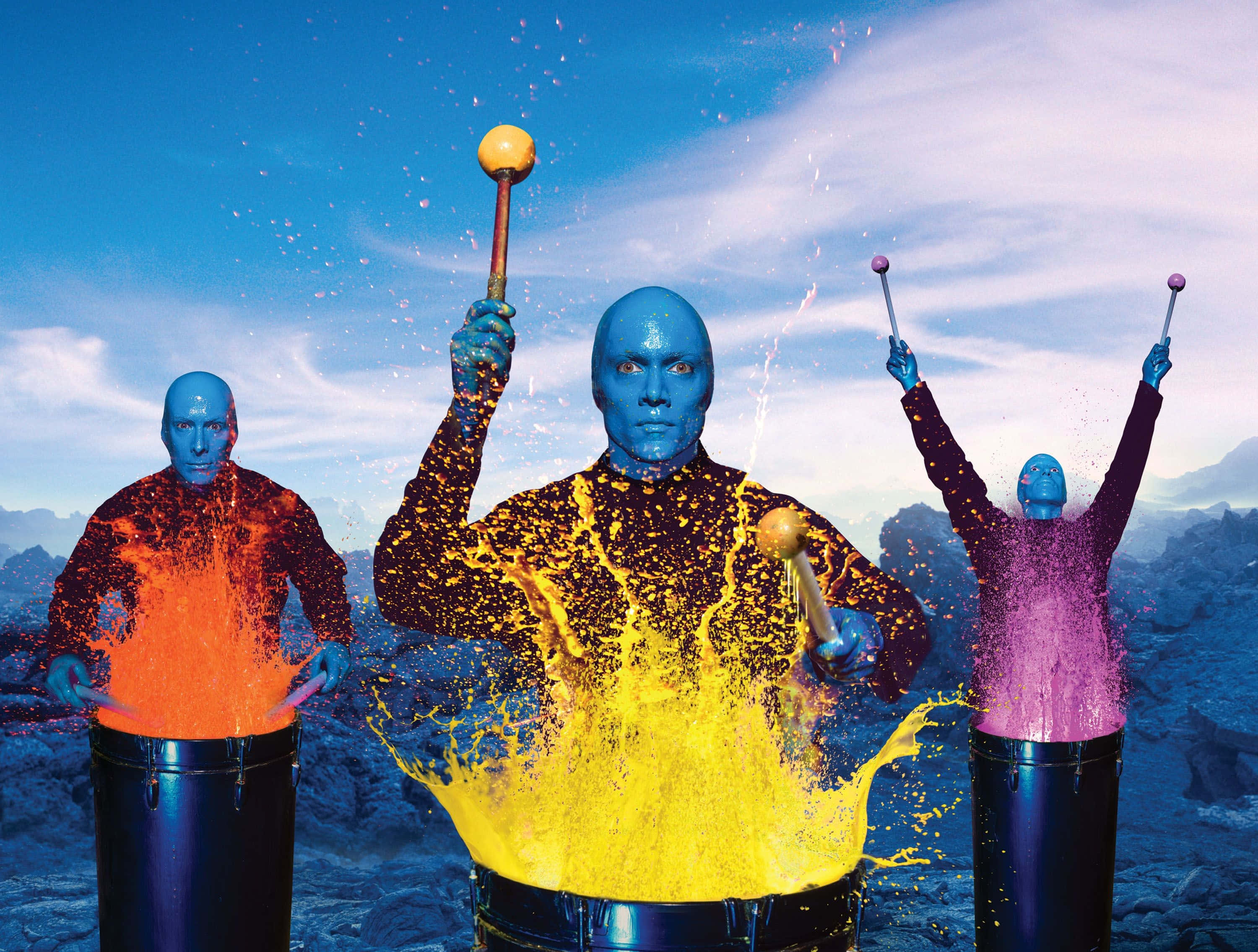 Blue Man Group 桌布 壁紙 — 三位藍皮膚的表演者在岩石景觀中擊鼓，鼓聲爆炸出鮮豔的橙色、黃色和紫色顏料飛濺，映襯著明亮的藍天。