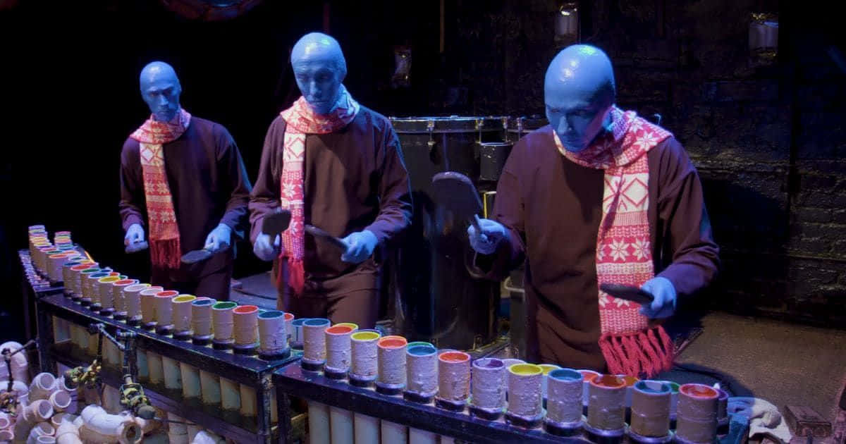 Blue Man Group 桌布 壁紙 — 三位藍皮膚表演者身穿棕色長袍和圖案圍巾，在昏暗的舞台燈光下演奏色彩繽紛的木琴。