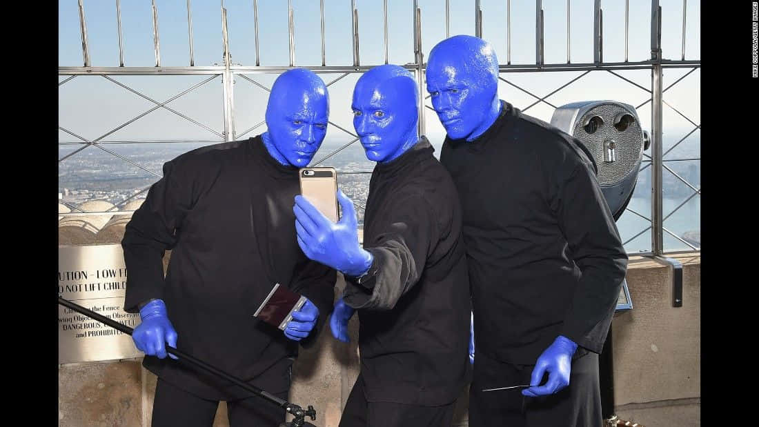 Blue Man Group 桌布 壁紙 — 三位藍臉表演者身穿黑色服裝，在高聳的觀景台上自拍，俯瞰廣闊的城市景觀。