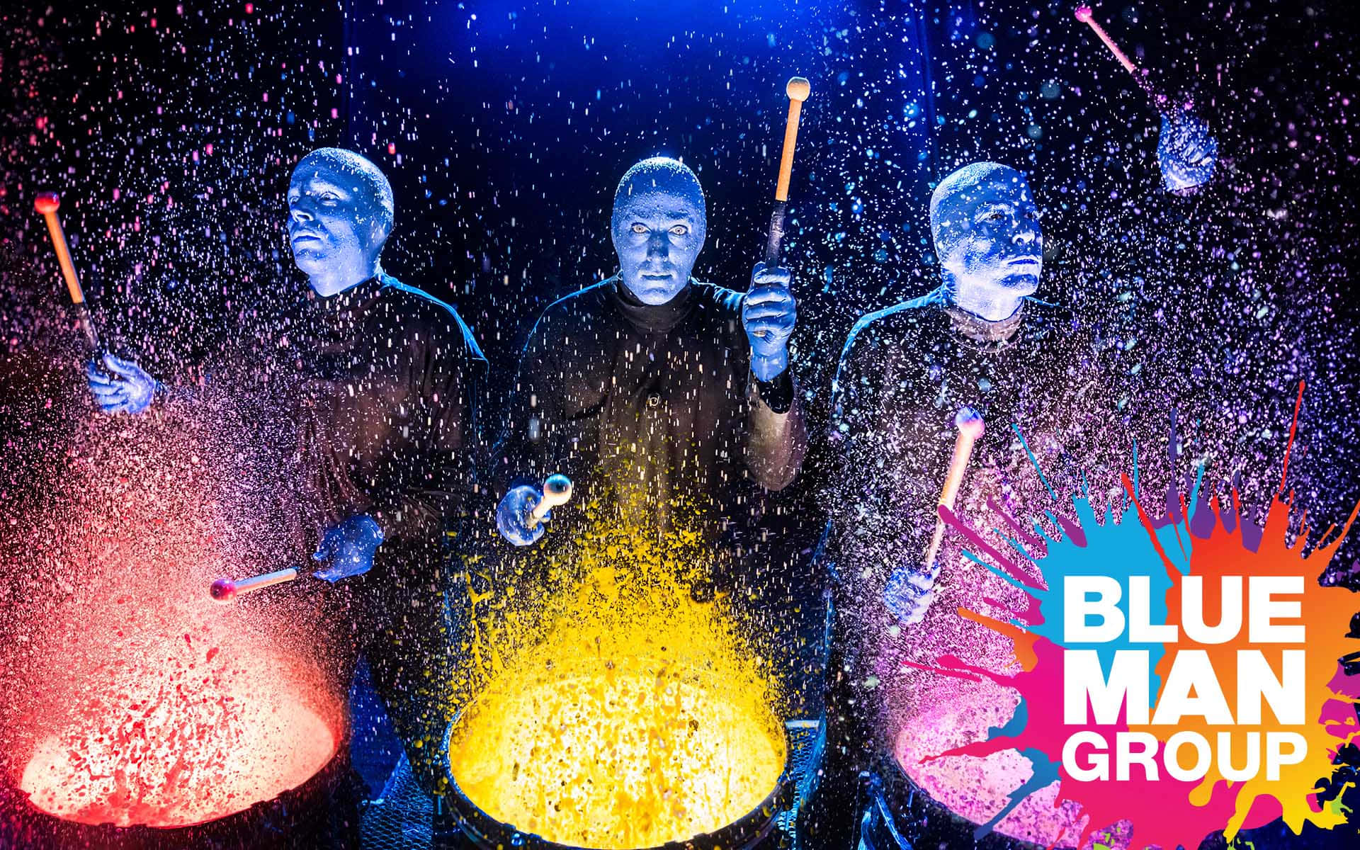 Blue Man Group 桌布 壁紙 — 三位藍面表演者在舞台燈光下，於色彩繽紛的顏料飛濺中充滿活力地敲打發光的鼓。