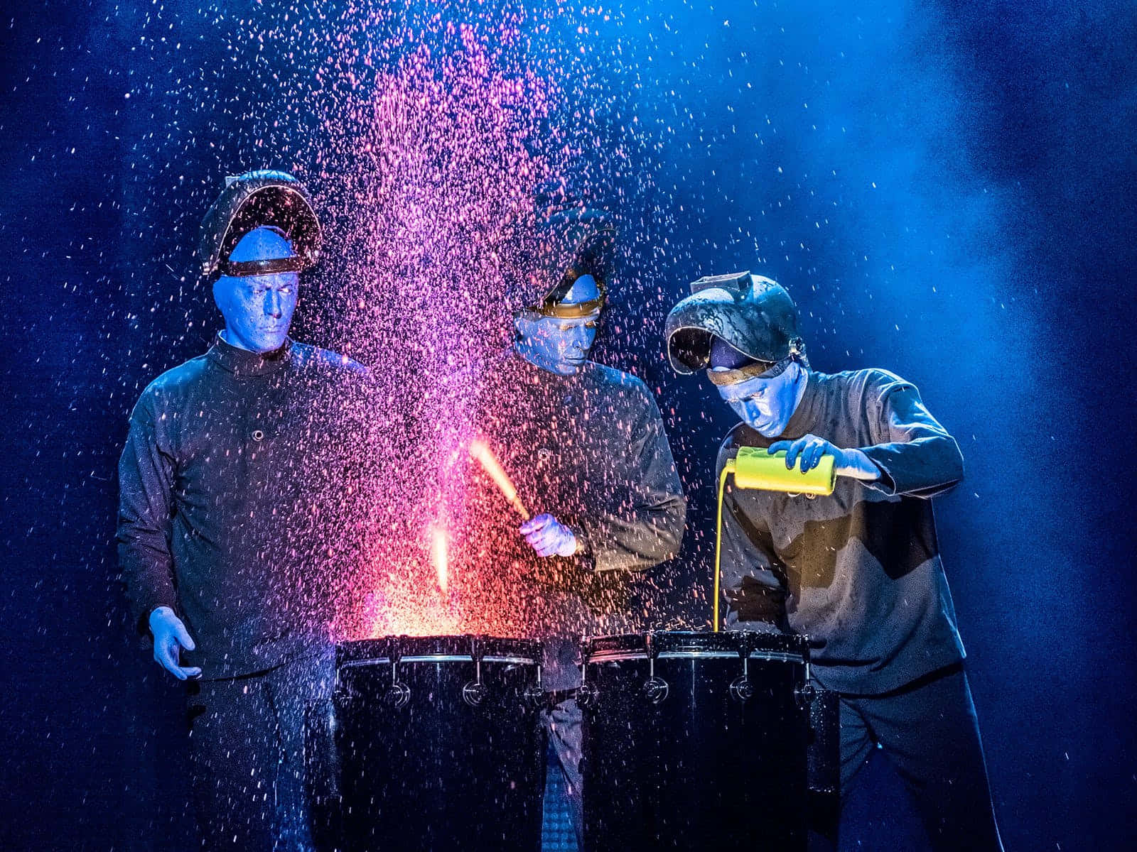 Blue Man Group 桌布 壁紙 — 三位藍面表演者戴著焊接頭盔，在戲劇性的藍色舞台燈光下，在鼓上創造出耀眼的粉紅色火花爆炸。