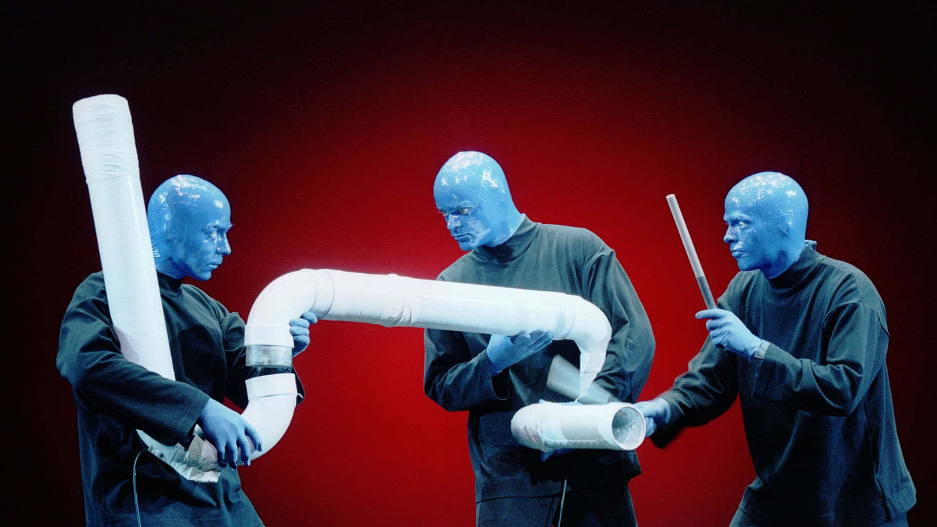Blue Man Group 桌布 壁紙 — 三位藍面表演者身穿深色衣服，在鮮紅色背景下操控白色PVC管。