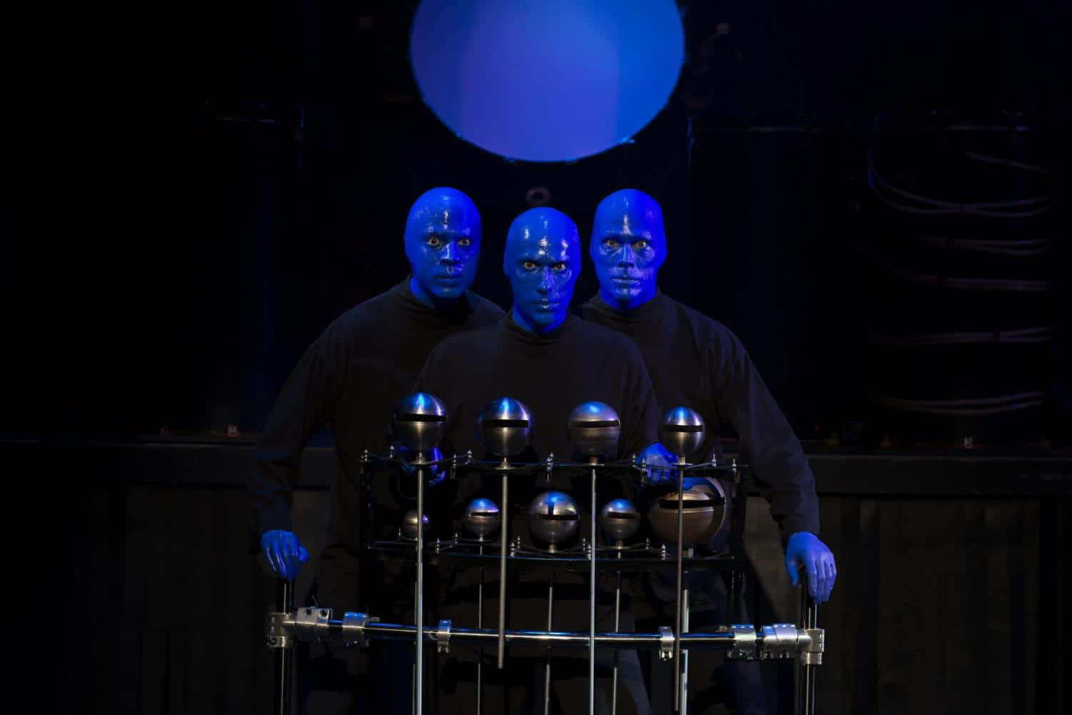Blue Man Group 桌布 壁紙 — 三位藍面表演者身穿黑色服裝，在黑暗中發光的藍色球體下，莊嚴地站在金屬敲擊樂器後面。