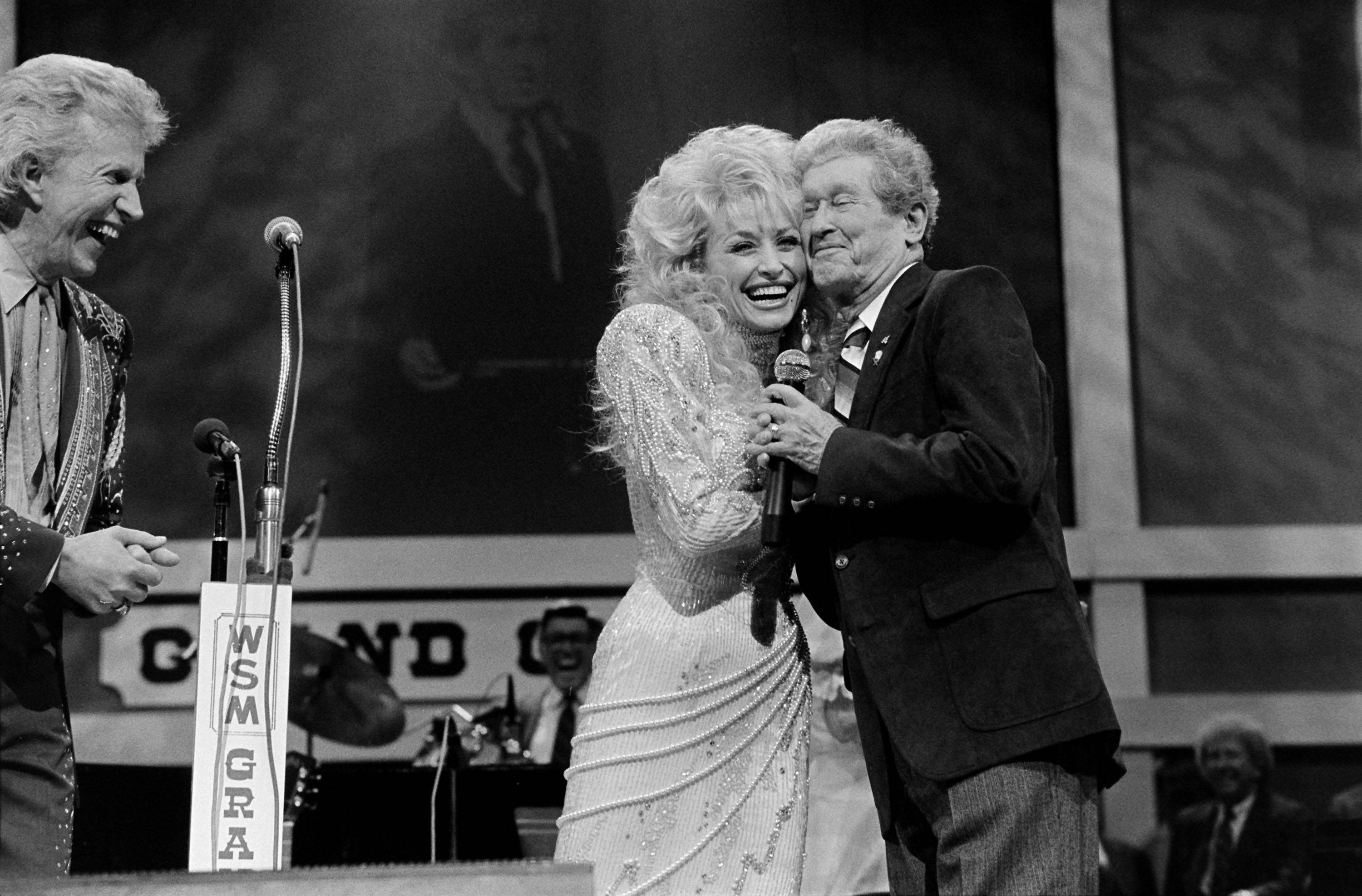 Dolly Parton 桌布 壁紙 — 三位表演者在舞台上分享歡樂時刻，身後有麥克風和「Grand Ole Opry」標誌。
