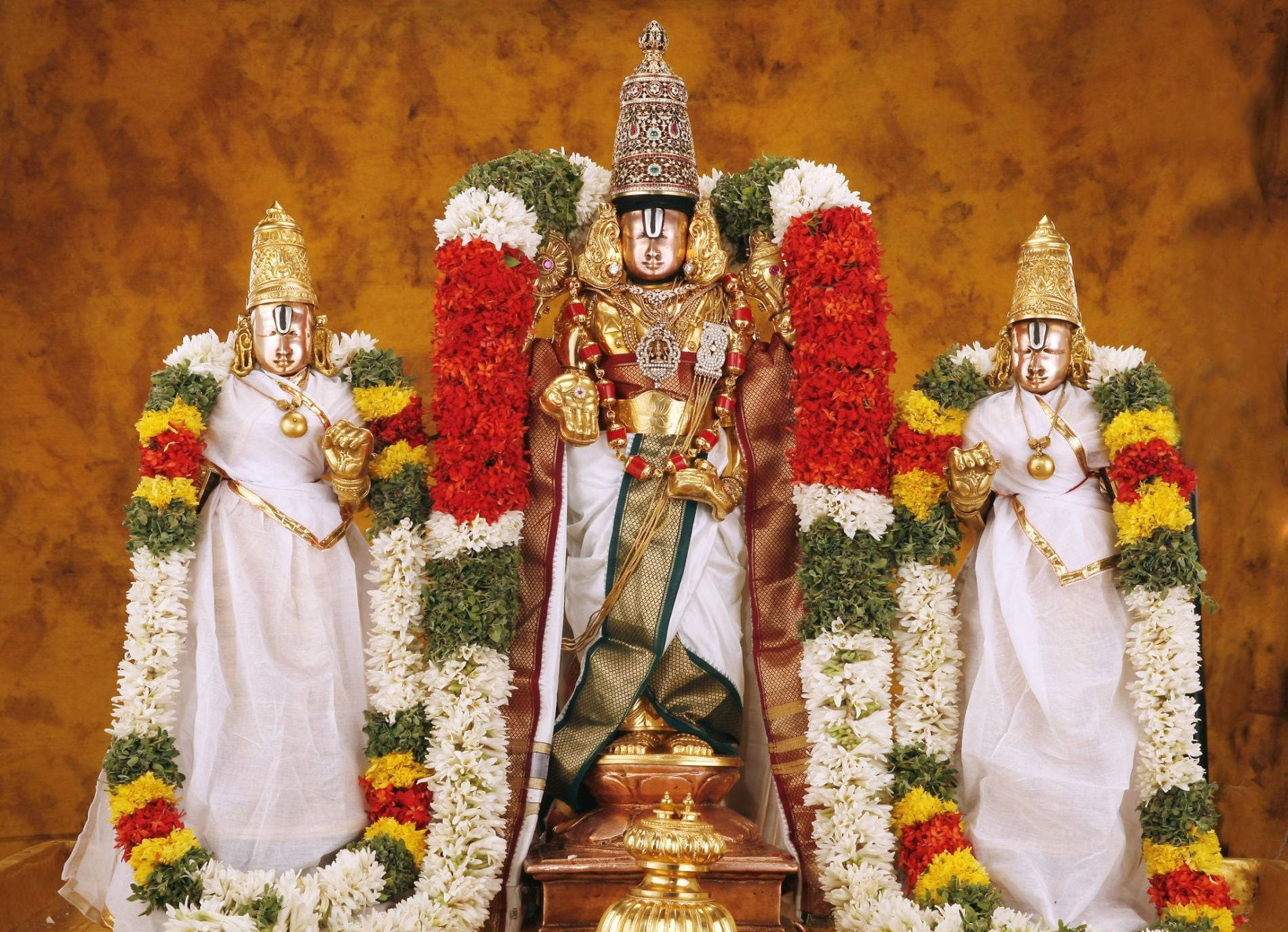 Venkateswara 桌布 壁紙 — 三位裝飾華麗的印度教神祇，佩戴著黃金珠寶和鮮豔的花環，莊嚴地站在溫暖的金色背景前。
