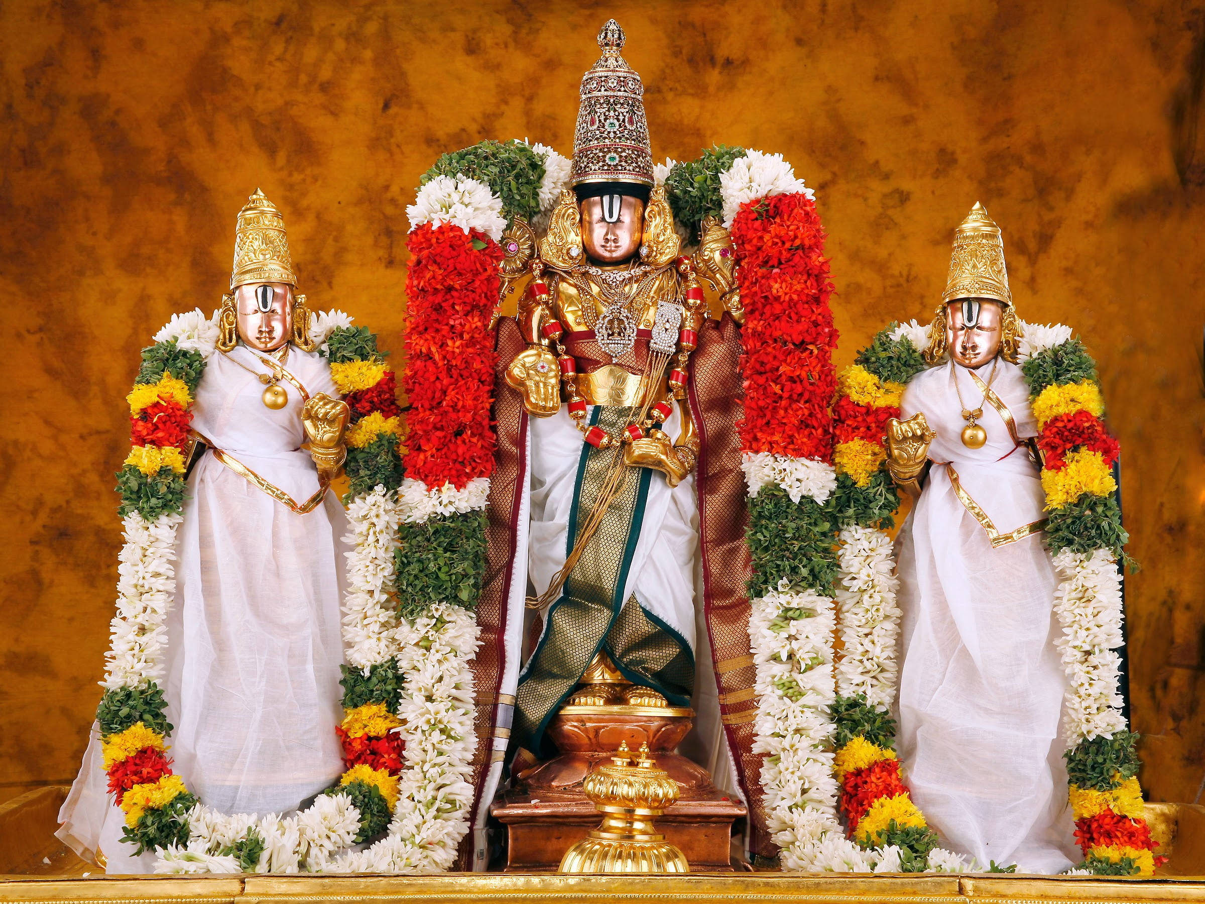 Lord Balaji 桌布 壁紙 — 三位裝飾華麗的印度教神祇，佩戴著黃金首飾和鮮豔的花環，莊嚴地站在溫暖的紋理背景前。