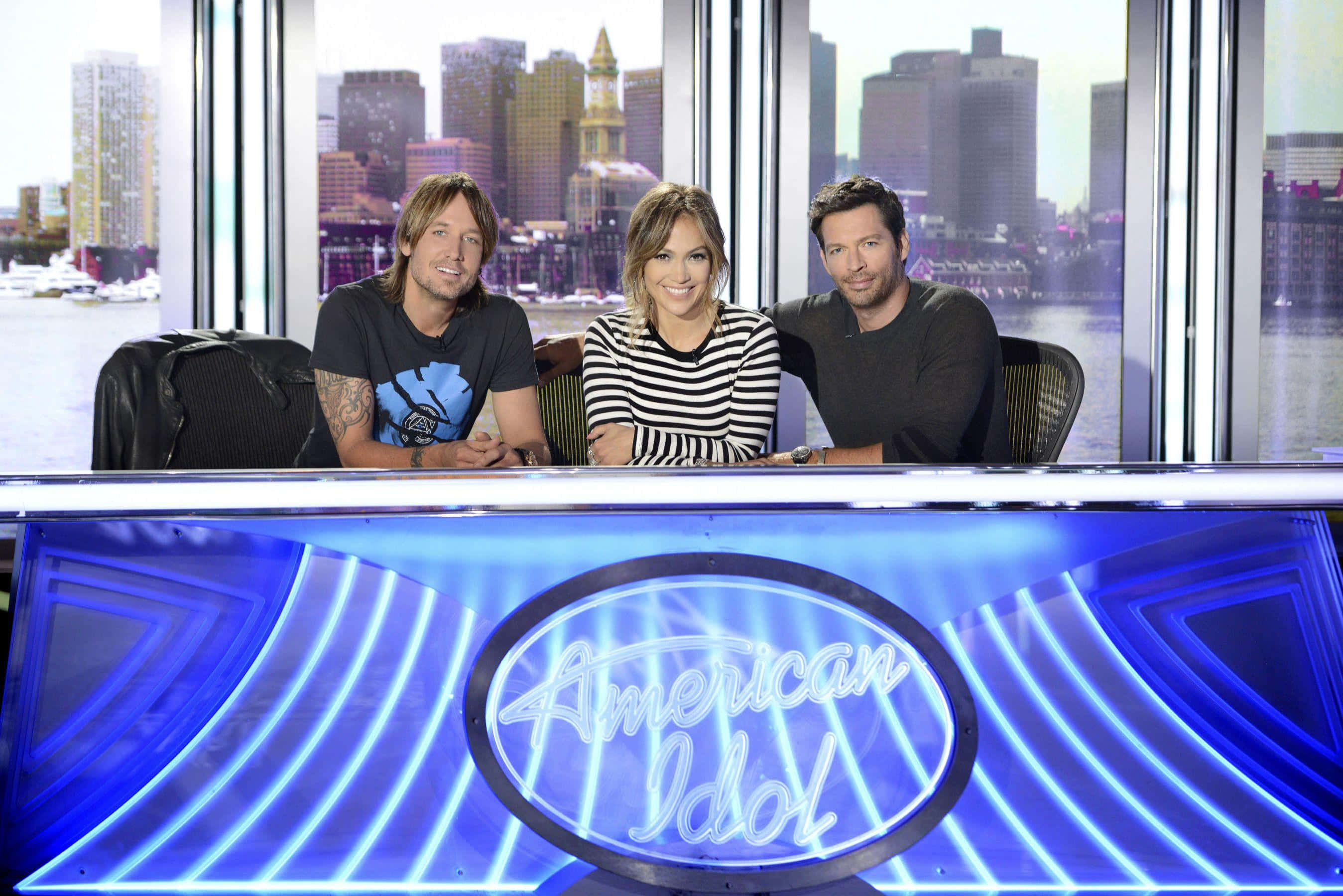 american idol 桌布 壁紙 — 三位評審坐在發光的藍色American Idol桌子後面，透過身後的大窗戶可以看到城市天際線。