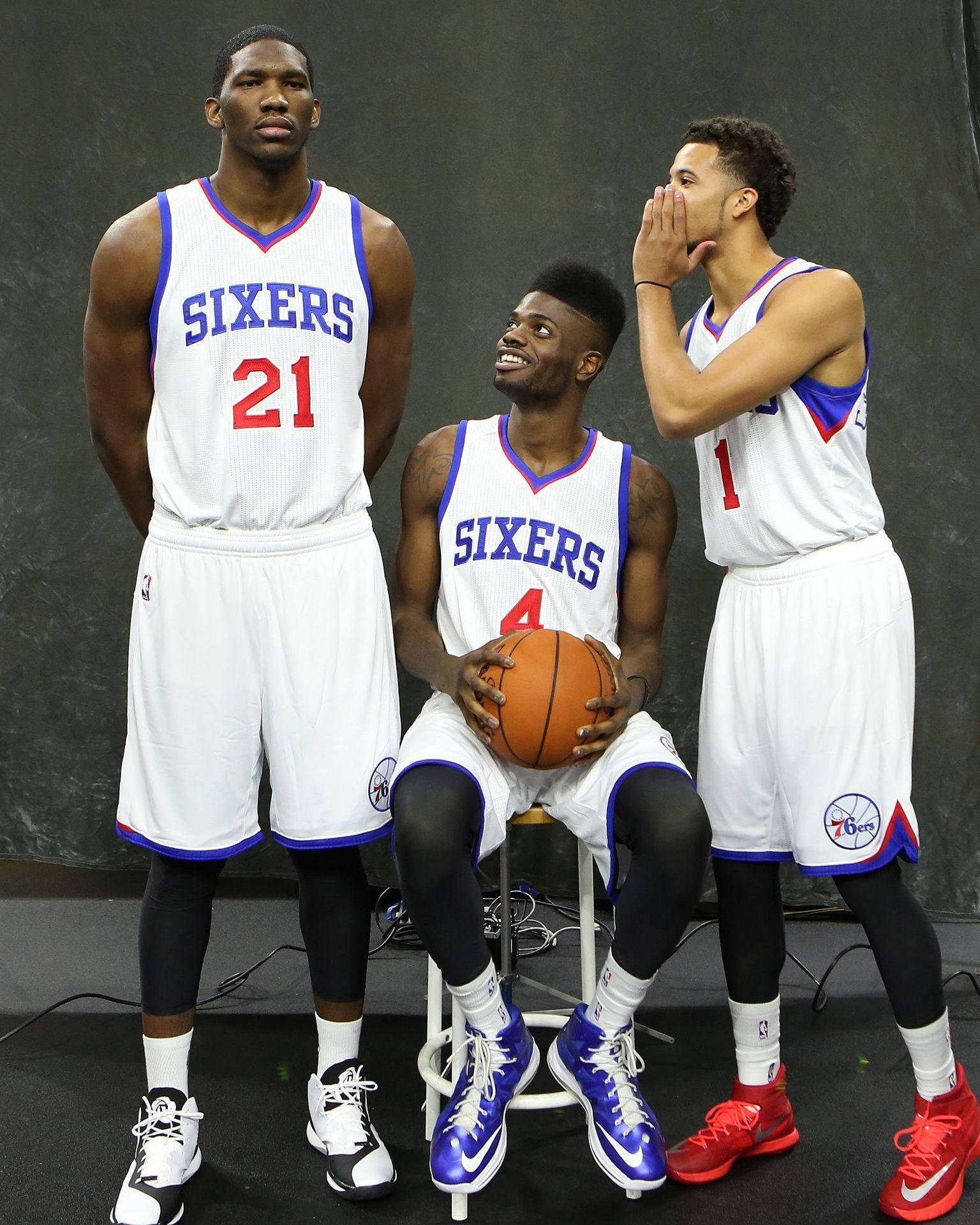 Embiid 桌布 壁紙 — 三位費城76人籃球運動員身穿白色球衣擺姿勢，一人坐著持球，另一人對他耳語。