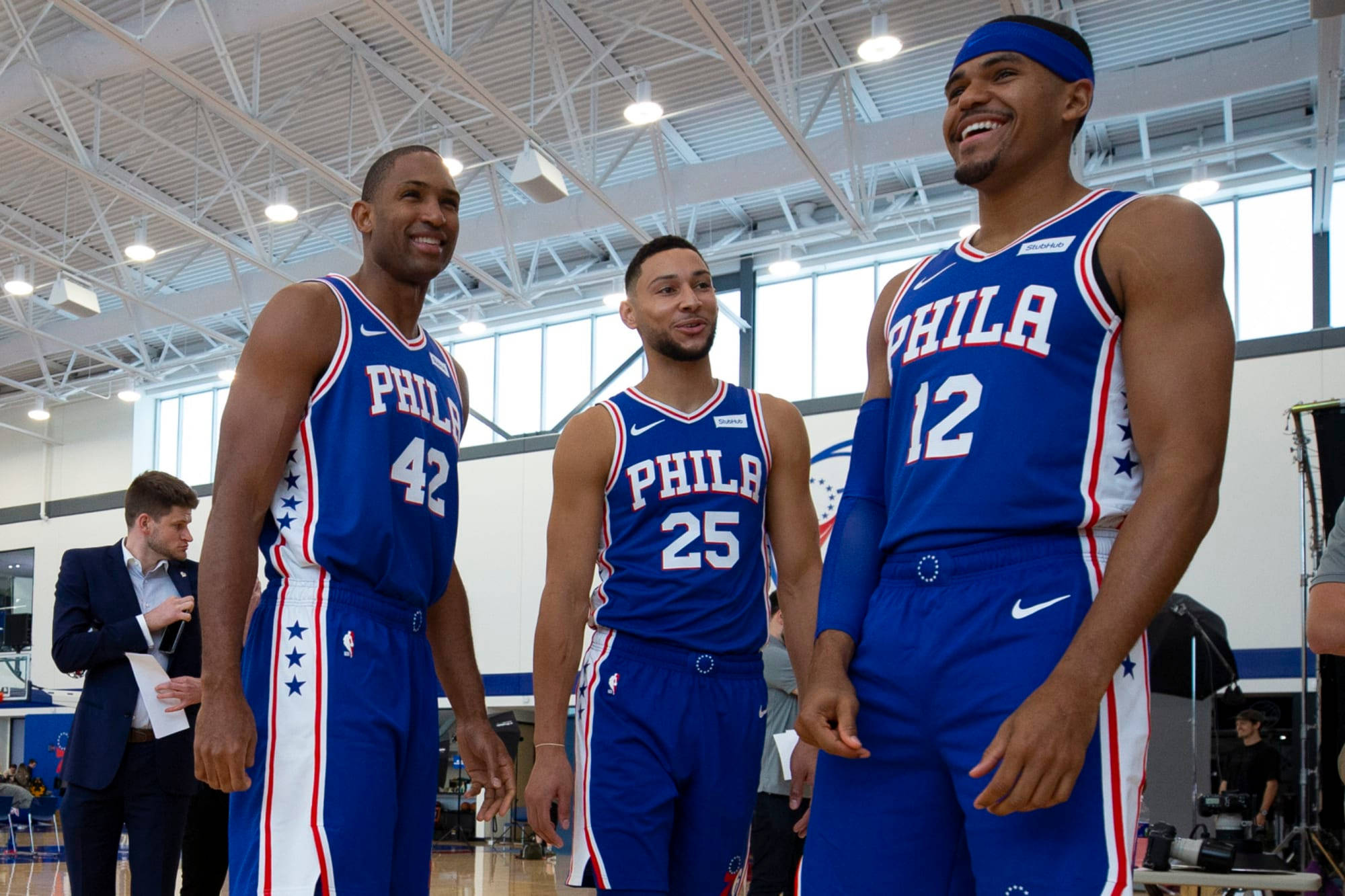 76ers 桌布 壁紙 — 三位費城76人籃球運動員身穿藍色星星圖案球衣，微笑著在明亮的室內體育館一起擺姿勢。