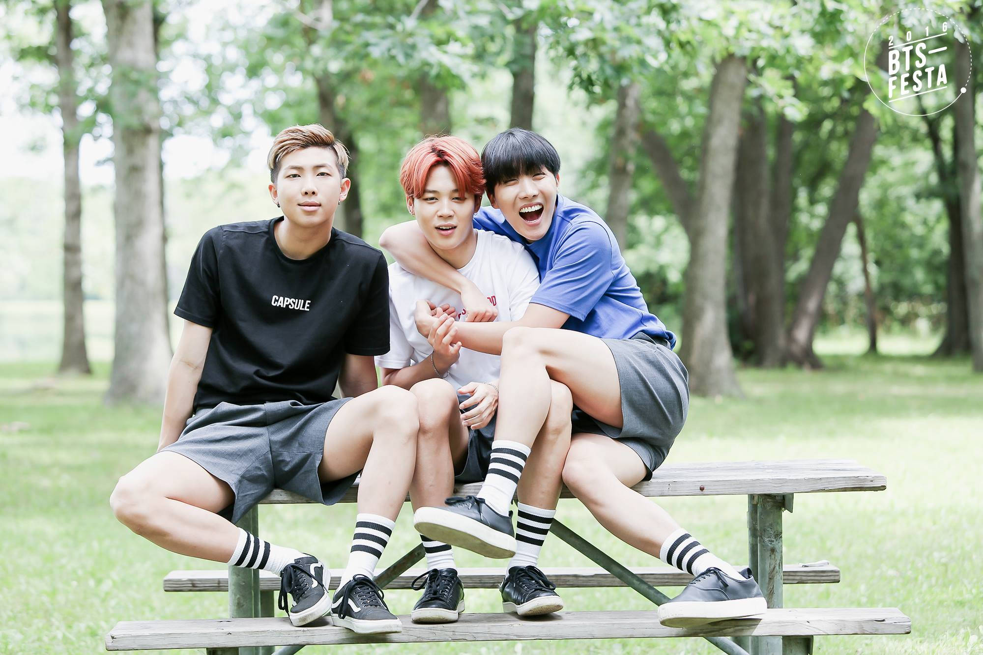 美學 BTS 桌布 壁紙 — 三位身穿休閒服的年輕男士在陽光明媚、樹木高聳的公園裡，一起坐在木製野餐長椅上大笑並擺姿勢。