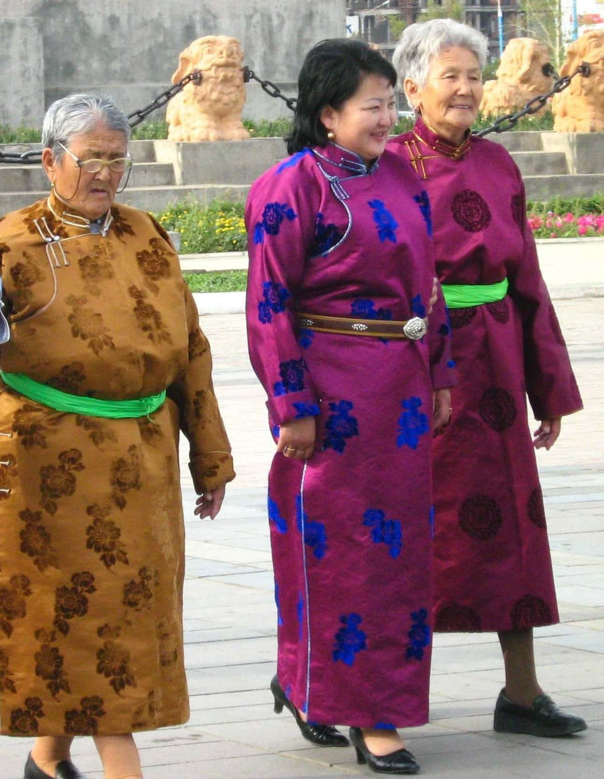 culture 桌布 壁紙 — 三位身穿傳統刺繡長袍的老年女性一起走在鋪砌的廣場上，背景有石獅雕像。