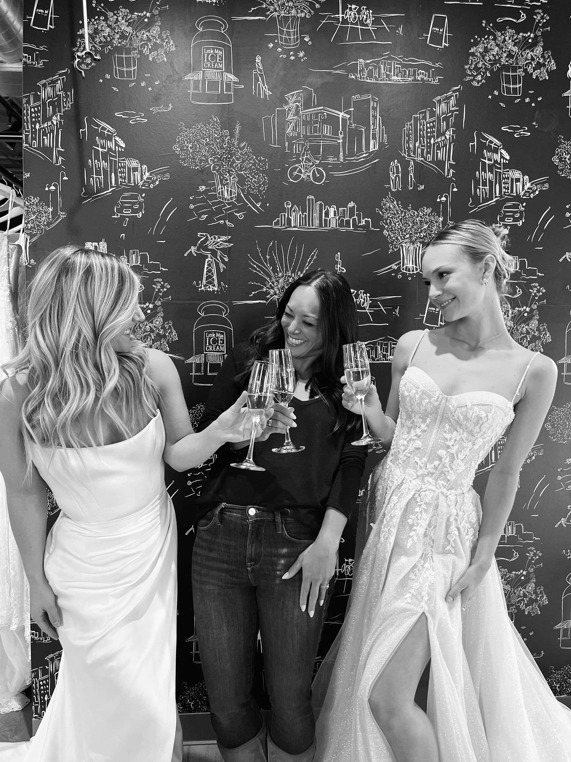 Bride 桌布 壁紙 — 三位身穿優雅禮服和休閒服裝的女性，在一個充滿奇思妙想的粉筆風格城市景觀牆前，用香檳杯乾杯。