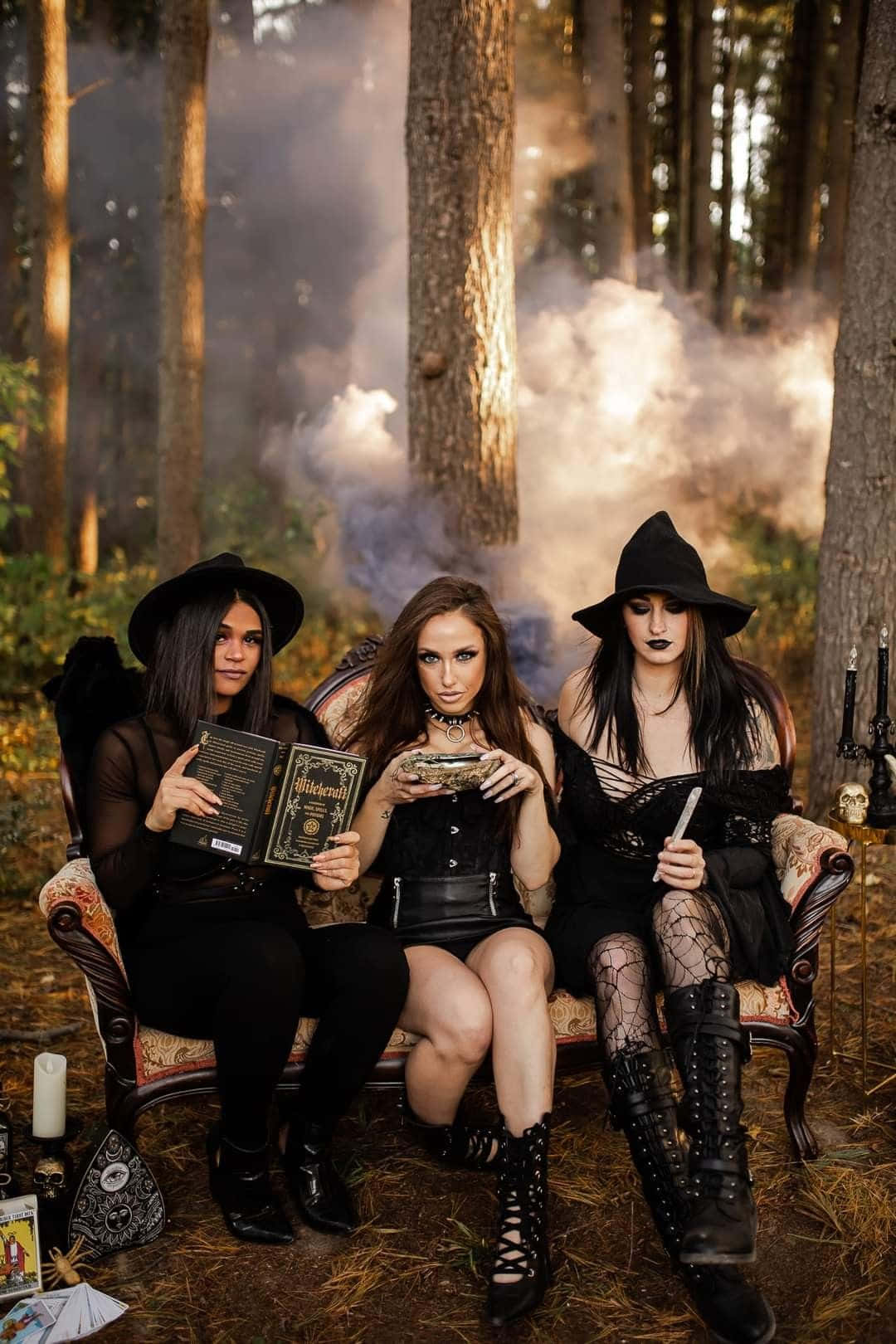Witch Costumes 桌布 壁紙 — 三位身穿女巫服飾的女性坐在煙霧瀰漫的森林中的華麗沙發上，手持魔法書和一個大鍋。