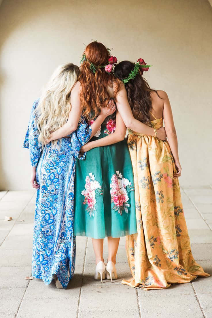 Bride 桌布 壁紙 — 三位身穿彩色花卉連衣裙的女性在瓷磚地板上從背後擁抱，每個人都戴著花冠。