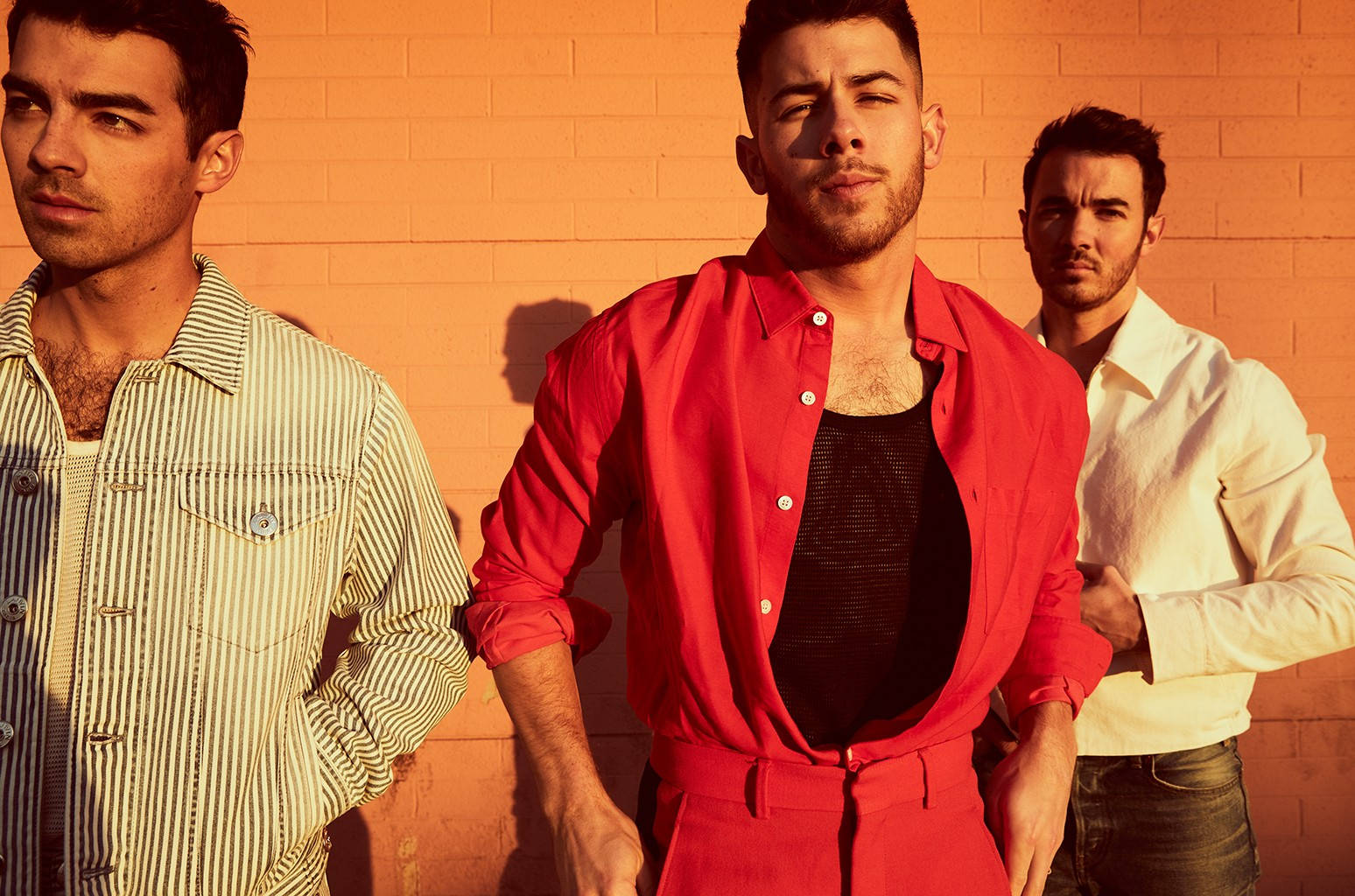 Jonas Brothers 桌布 壁紙 — 三位身穿時尚服裝的男士在金色時光的照耀下，自信地站在溫暖的橙色磚牆前。
