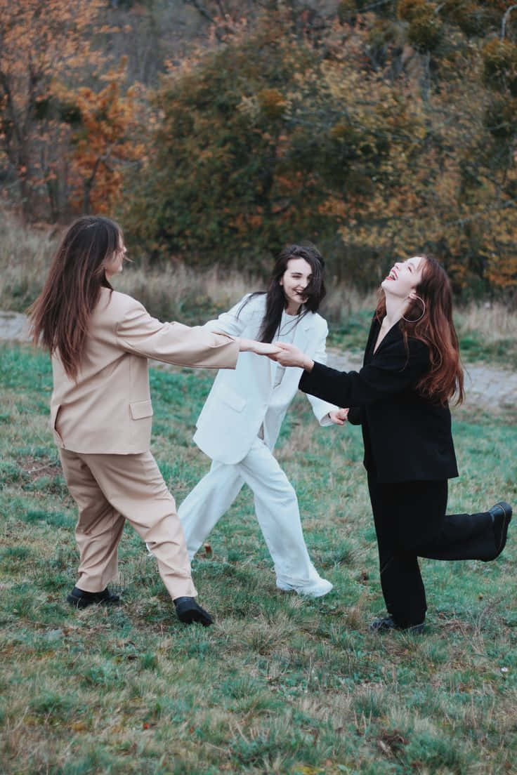Three Friends 桌布 壁紙 — 三位身穿時尚西裝的女性在秋季的草地上愉快地互相追逐嬉戲。