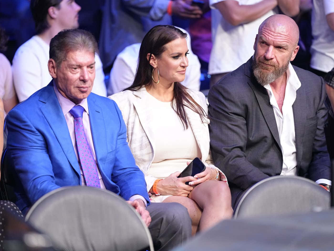 WWE 桌布 壁紙 — 三位身穿正裝的觀眾在燈火通明的活動中坐在體育場座位上，神情嚴肅地專注前方。