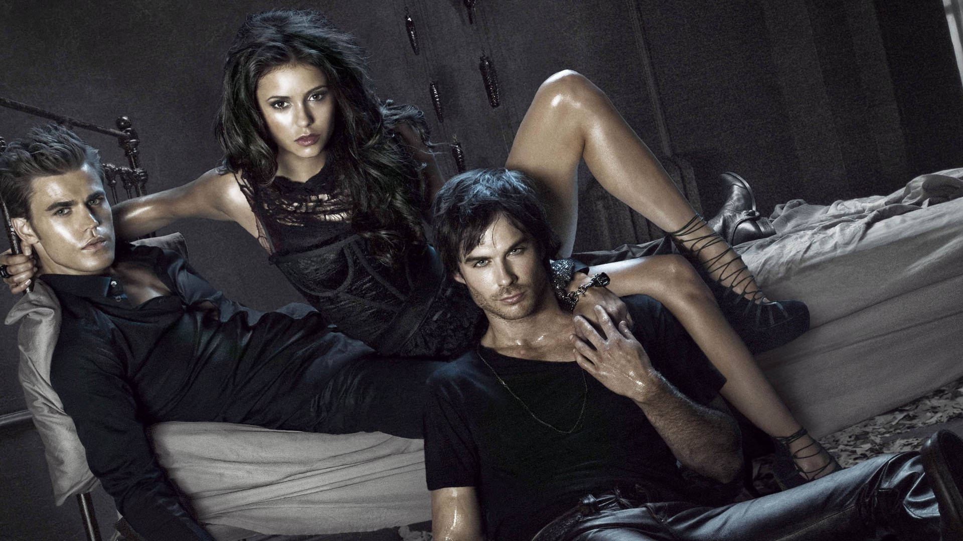 The Vampire Diaries 桌布 壁紙 — 三位身穿深色服裝的演員在昏暗、氣氛憂鬱的房間裡的床上，背景有武器，擺出強烈的姿勢。
